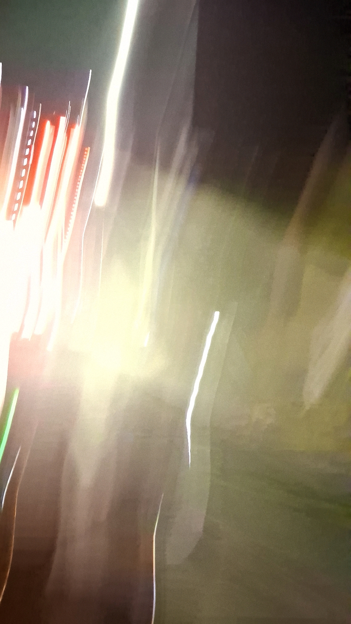 This is an abstract, motion-blurred photograph taken at night in Berbah, Indonesia. The image is dominated by streaks of light, primarily in white and orange hues, suggesting moving vehicles or streetlights. The background is dark with hints of green and brown, possibly indicating foliage or a dimly lit outdoor environment. The blur effect creates a sense of movement and dynamism, making it difficult to discern specific objects or subjects within the frame. There are no identifiable people or clear structures. The overall impression is one of urban nighttime activity captured with intentional camera movement.
