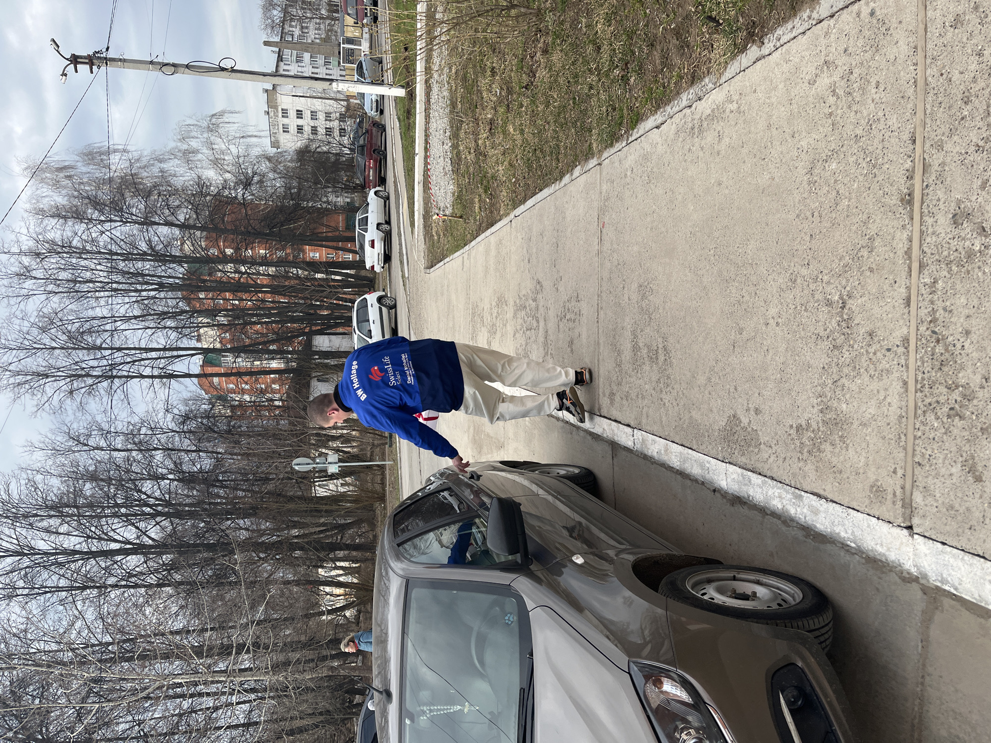 The image depicts an urban street scene in Chaikovsky, Russia, on an overcast day, likely during late winter or early spring due to the bare trees.
In the foreground, a man in a blue jacket and light-colored pants is opening the driver's side door of a dark grey or brown compact car. The back of his blue jacket displays white text and a logo, including phrases like "BW Holidays," "SwissLife," and "Official Representative," along with a red, stylized emblem that resembles a hot air balloon or an 'S'. A partial reflection of another person is faintly visible in the passenger-side window of the car.
The car is parked on a paved surface adjacent to a concrete sidewalk. Beyond the sidewalk, a narrow strip of patchy, dry grass runs alongside a row of tall, bare deciduous trees. In the background, several multi-story apartment buildings are visible through and above the tree line. A concrete utility pole with electrical lines and fixtures stands prominently against the grey sky on the left side of the frame. Further down the paved area, other cars, including white and dark-colored models, are parked.
The scene captures a routine moment in a residential or mixed-use urban environment, characterized by functional architecture and early seasonal foliage.