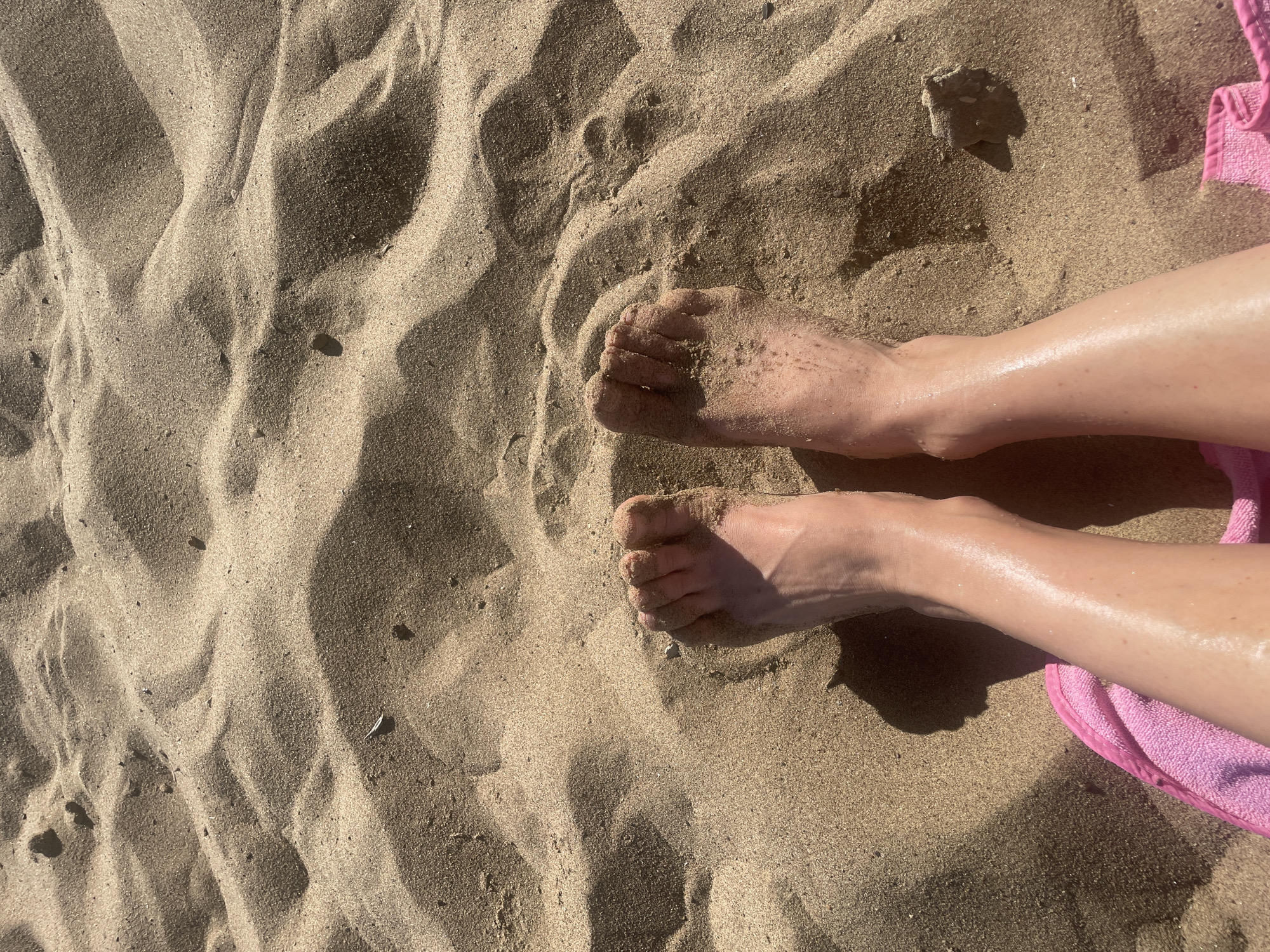 The image shows a pair of human legs and feet resting on a sandy surface, likely a beach. The feet are partially covered in sand. Next to the legs is a pink towel. The sand appears to be textured with dunes. It is unlikely that this image was taken in the city of Saratov, Russia, which is located far inland.