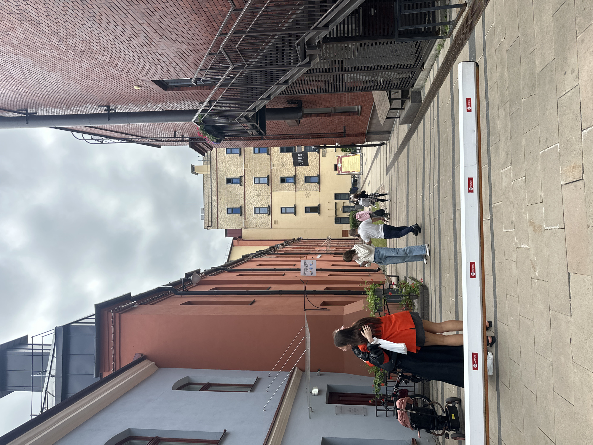 The image depicts an outdoor scene in an alleyway or courtyard, likely within a city. The environment features several buildings with distinct architectural styles. On the left, a pale blue building with arched windows and cream-colored trim is visible. Adjacent to it is a salmon-colored building with a darker section at the top. The right side of the alley is dominated by a red brick building with an external metal staircase leading upwards. Further down the alley, a tall, light-colored building with multiple windows is present. The ground is paved with rectangular stone tiles.
In the foreground, a white barrier with red circular logos is positioned horizontally across the walkway. Behind this barrier, two individuals are embracing; one is wearing a red jacket and a black skirt, and the other is wearing a black outfit. Several other people are walking in the background, some looking at their phones. There's also a stroller visible on the left side of the image.
The sky is overcast with grey clouds. The overall impression is of a pedestrian area within an urban setting. The presence of a stroller and people casually dressed suggests it might be a public space, possibly a commercial or residential area.
The location is in the city of Minsk, Belarus. The specific location within Minsk is the courtyard of the "Oktyabrskaya" creative hub, also known as the former Oktyabrskaya factory. This hub is situated on Oktyabrskaya Street, a well-known area in Minsk for its revitalized industrial spaces that now house various cultural and business establishments.