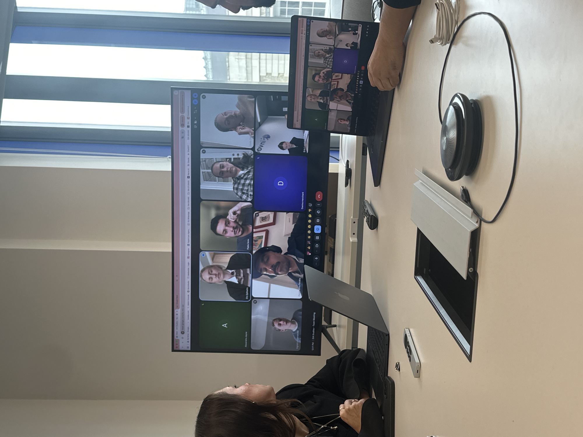 The image depicts a meeting taking place in a well-lit office space. The primary focus is on two screens displaying a video conference call with multiple participants. On the larger screen, several individuals are visible in individual video feeds, including people named "Alexandra Jayne," "Elin Sjöholm," "Pablo Soto," "Davina merchant," and "Eric Lunds." Additionally, logos for "SOUNDSTUDIO Cort" and "OVNI" are visible within the video feeds. A second laptop screen displays a similar video conference layout.
At least two people are present in the room. One woman is seated in front of one laptop, and the hands of another person are visible near the second laptop.
Additional objects present on the table include a speakerphone with its connecting cable, a covered power outlet, and a smartphone.
The environment is a modern office with a large window. The window gives a glimpse of an urban landscape outside, potentially suggesting a location in a city such as London, United Kingdom. The presence of modern office equipment suggests a professional workplace.