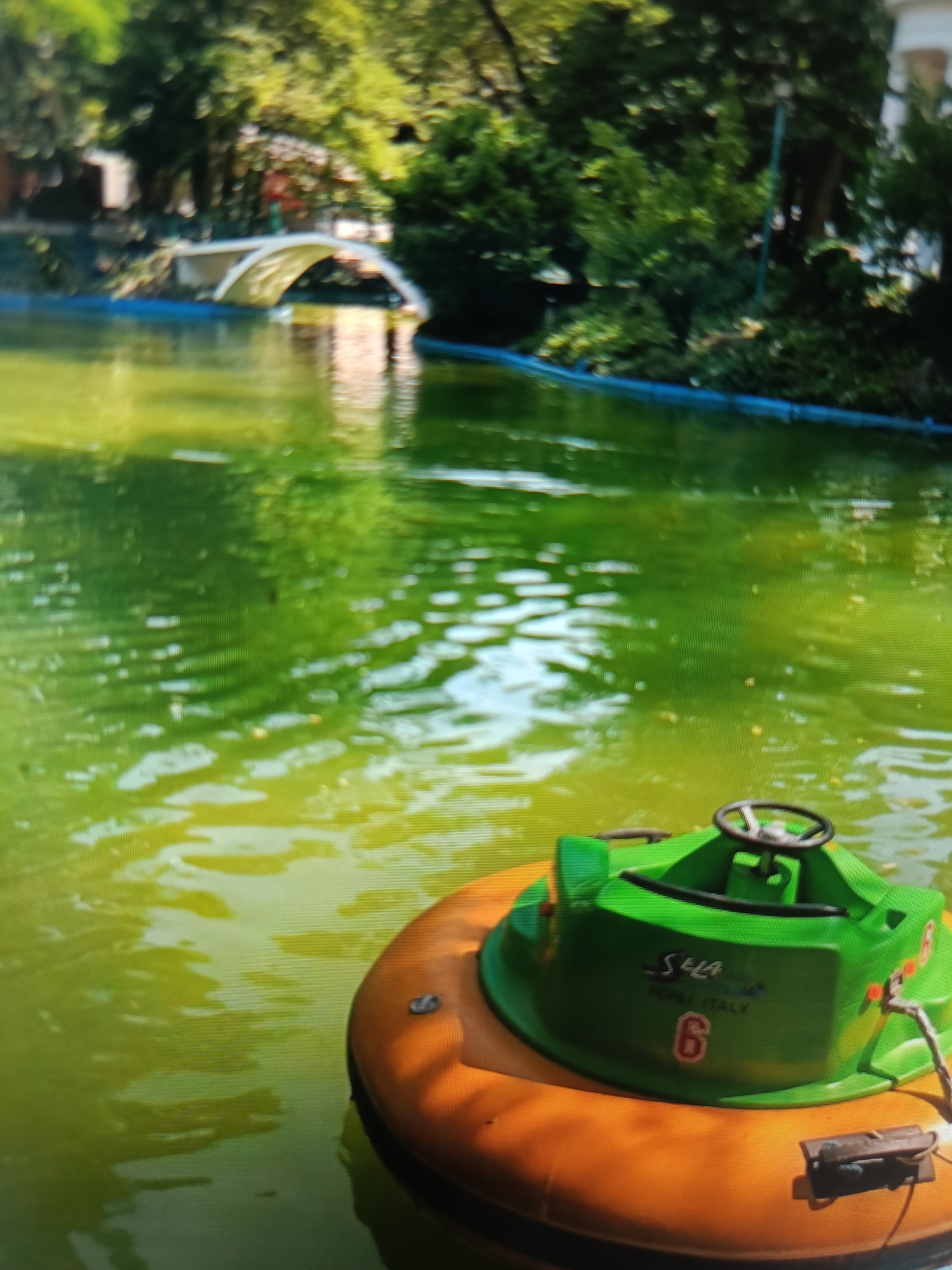 The image depicts a small, orange bumper boat with a green top, a steering wheel, and the number "6" visible on its side, floating on a body of green water. In the background, a white arched bridge spans across the water, surrounded by lush green trees. The scene appears to be a park or recreational area. The image does not provide any specific clues to suggest it is located in Varna, Bulgaria.