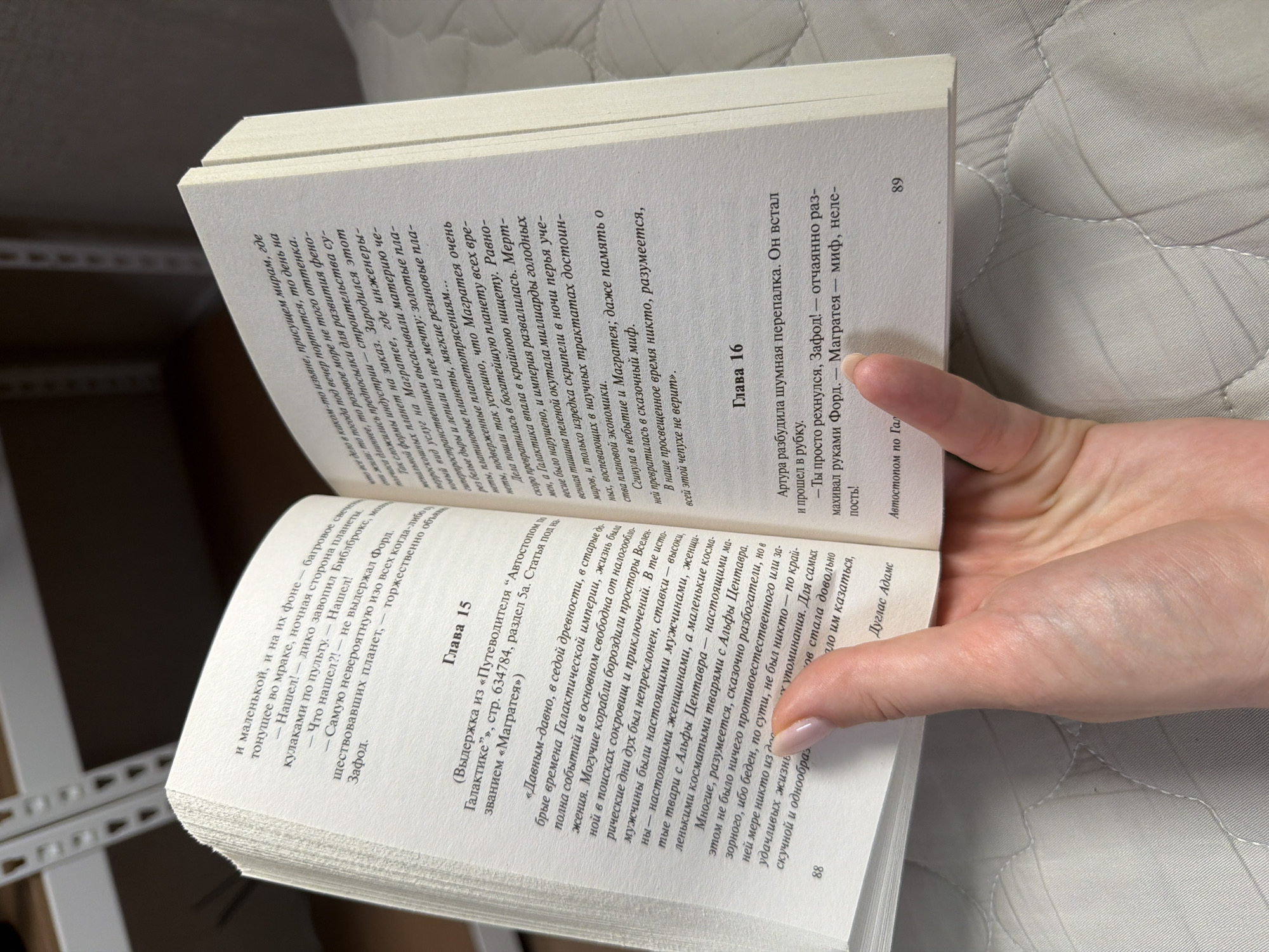 An open book is held by a person's hand in a close-up shot. The book is in Russian, with text visible on both pages. The left page has a chapter title, "Глава 15" (Chapter 15), and a page number, "88." The right page has the chapter title "Глава 16" (Chapter 16) and the page number "89." The person's fingernails are painted with a light pink polish. The background is blurry and appears to be a quilted surface. The lighting is soft and natural. There are no other people or objects clearly visible in the image. The location in Dongducheon-si, South Korea is not discernible from the image.