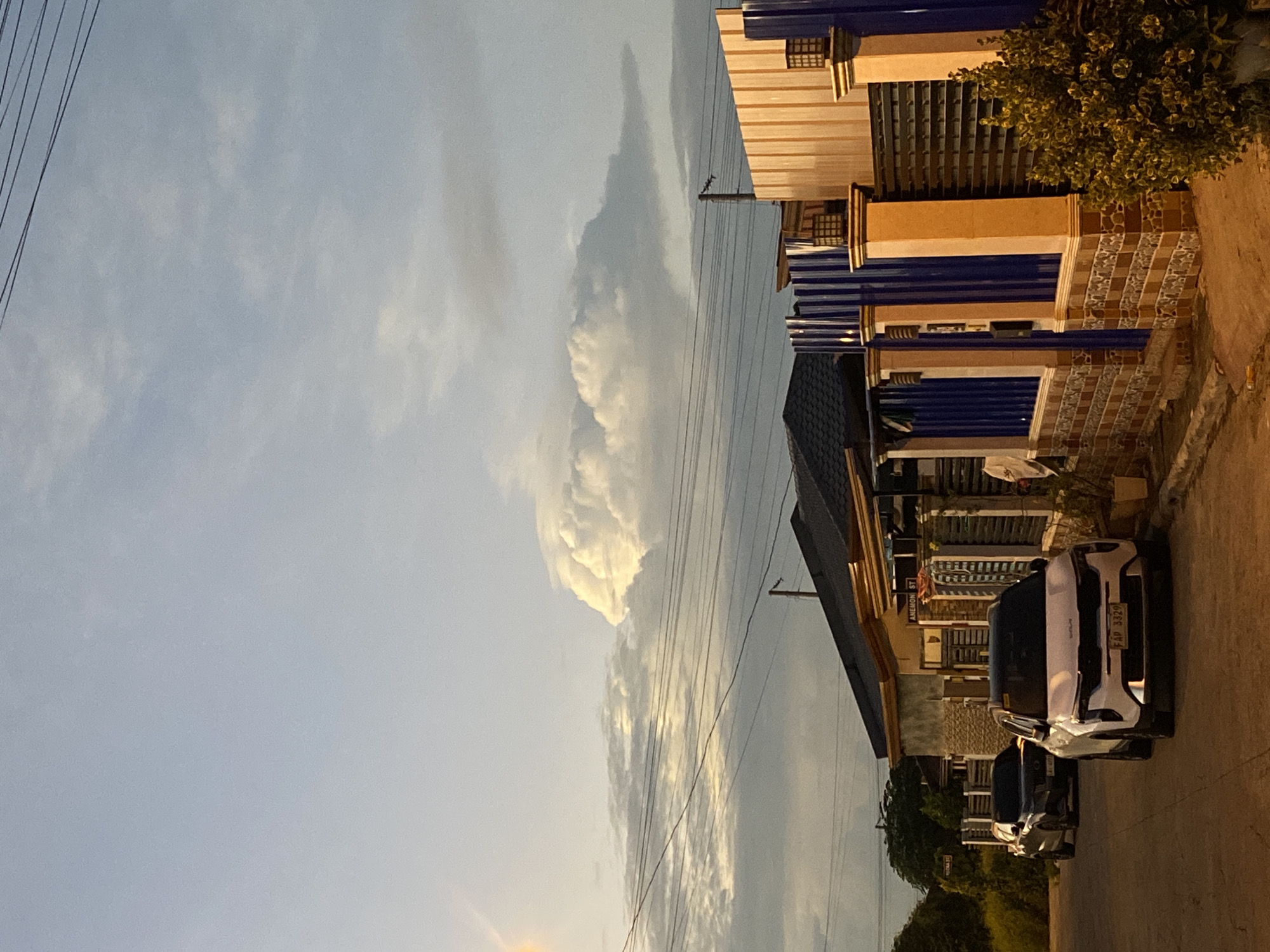 The image depicts a residential street scene in Davao City, Philippines, during what appears to be either early morning or late afternoon, judging by the soft lighting and sky conditions. Two vehicles are parked on the paved street. A white SUV, a Kia model, is in the foreground, facing forward with its license plate visible as "FAP 3329". Behind it, a dark-colored SUV is also parked. The street is lined with fences and houses, one of which features a sign indicating "ANEMONE ST". The architecture of the houses appears to be modern residential style, with tiled roofs and decorative fencing. Overhead, power lines crisscross the sky. The sky itself is a prominent feature, with a large, fluffy cumulus cloud formation dominating the upper portion of the frame, illuminated by the sun, creating a dramatic contrast with the softer, layered clouds on the horizon. The overall impression is a quiet, suburban street.