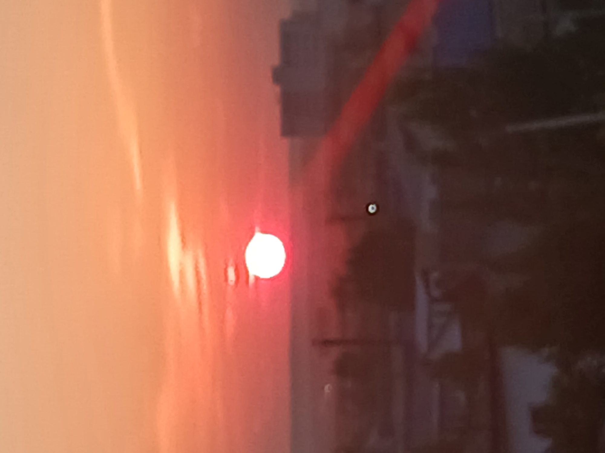The image depicts a sunset over the city of Makinsk, Kazakhstan. The sun, a bright orb, is positioned in the center of the frame, casting a warm, orange glow across the sky. Wispy clouds are visible, painted in shades of pink and orange. In the background, the blurred silhouettes of buildings and trees suggest an urban landscape. A lens flare is present on the right side of the image, appearing as a red streak. The overall atmosphere is serene and tranquil, capturing the beauty of a sunset in a distant city.