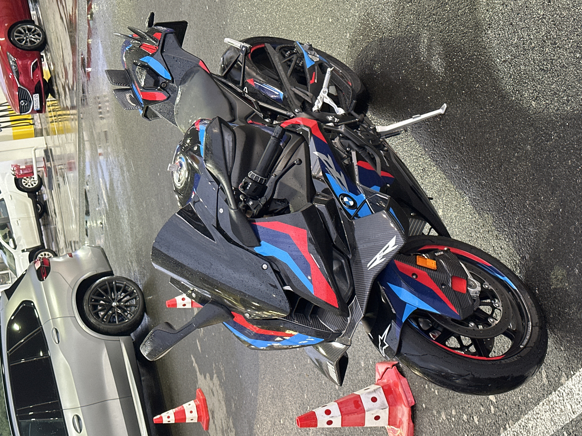 This image depicts a black BMW sport motorcycle with a distinctive red, blue, and white graphic design parked on wet asphalt. The motorcycle is positioned in the foreground, angled towards the right side of the frame. It features carbon fiber accents, a raised rear section with small winglets, and a low-slung front fairing with dual headlights. In the background, several cars are visible, including a silver SUV, a white sedan, and a red Mazda. Orange and white traffic cones are placed around the motorcycle. The environment appears to be an outdoor parking area or lot, possibly at a gas station or car wash, given the wet surface and the presence of vehicles that suggest a public thoroughfare or commercial establishment. The lighting suggests daylight, with reflections visible on the wet pavement. The scene is set in Drazhevo, Bulgaria.