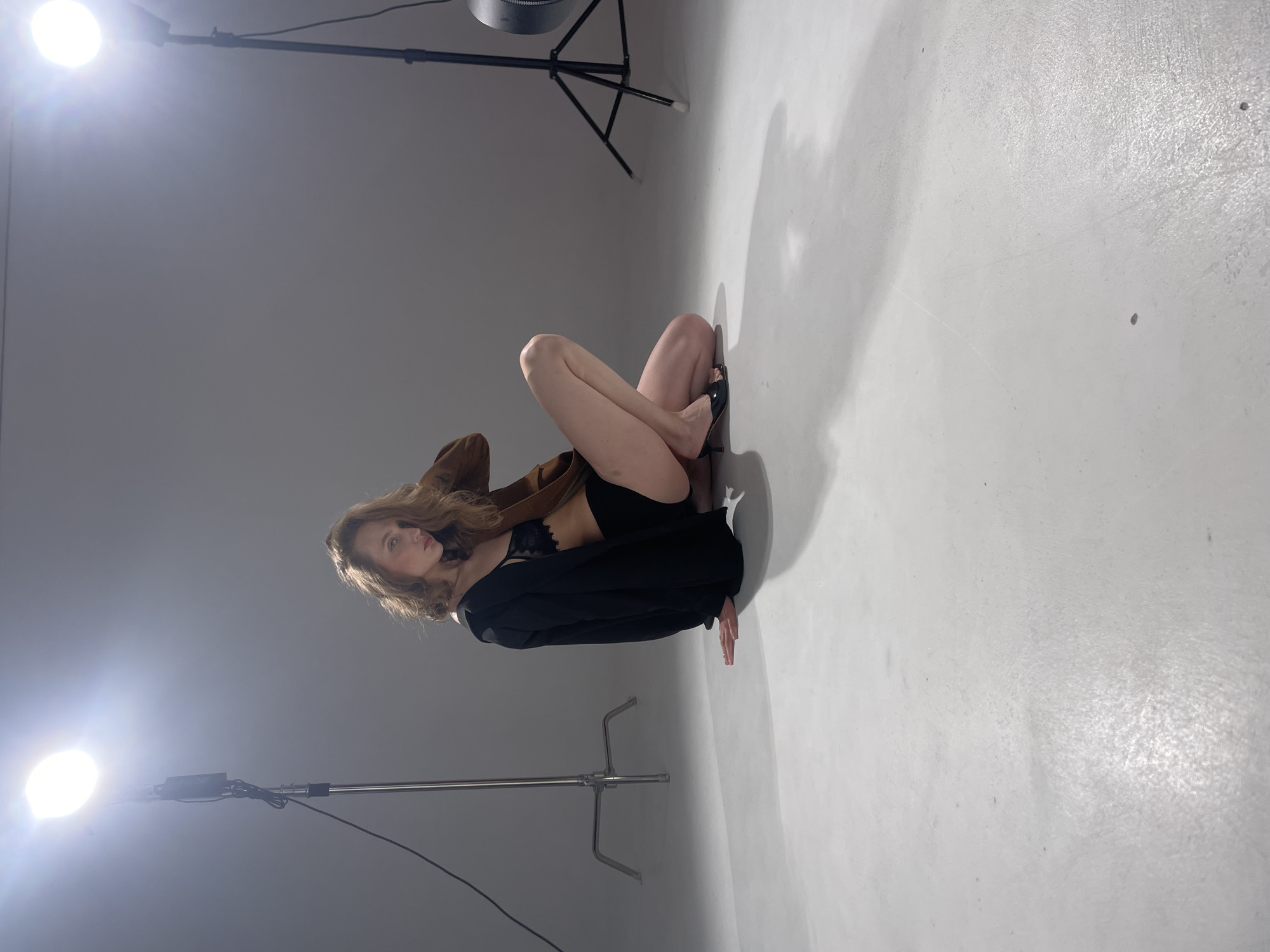 A woman is crouching on a white surface in what appears to be a photography studio. She is wearing black underwear, a brown jacket, and a black blazer. She is also wearing black heels. The woman has curly blonde hair and is looking at the camera. There are two light stands in the scene, one on each side of the woman. The location is not evident from the image.
