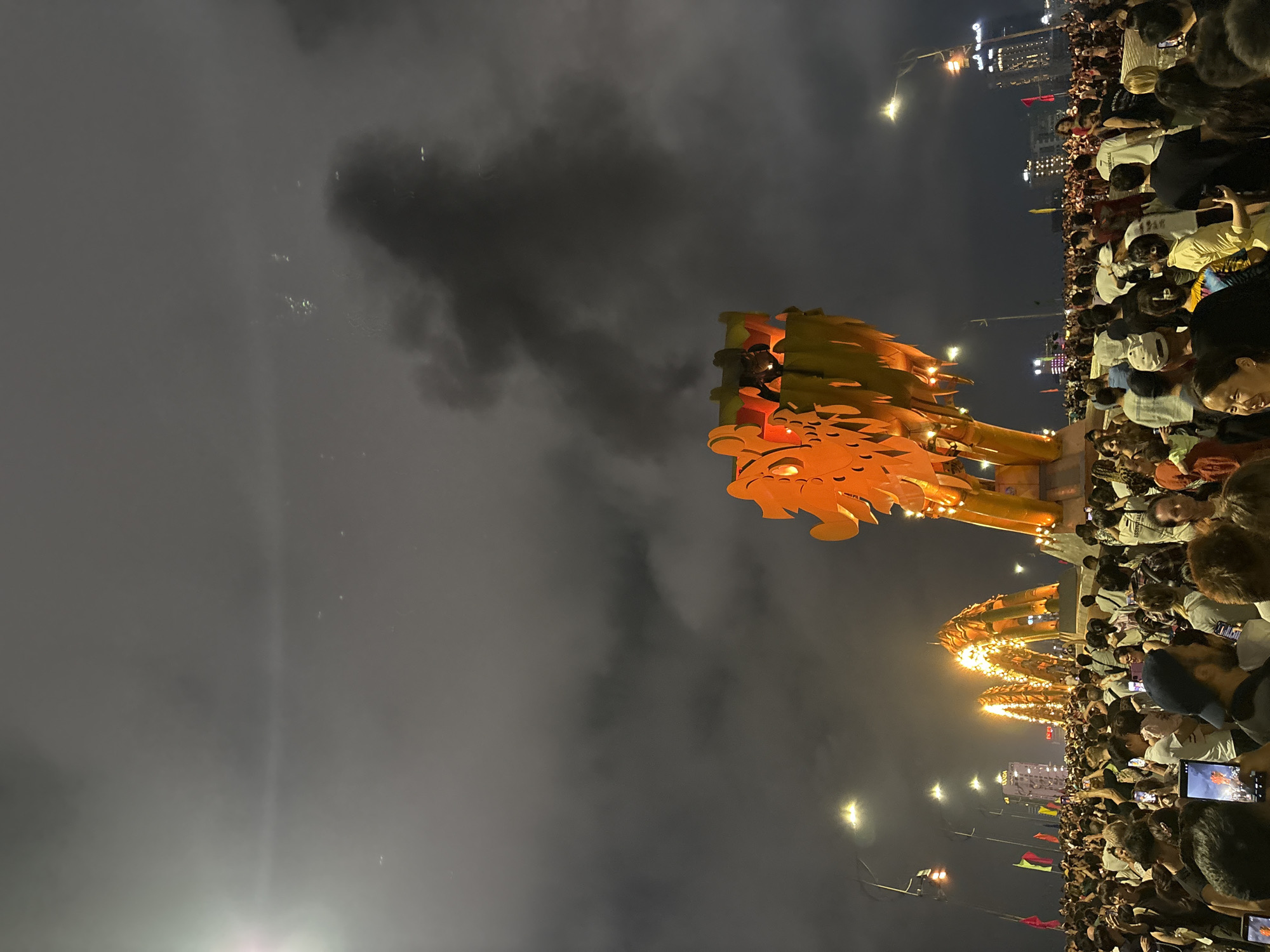 A vertical shot captures a nighttime scene in Đà Nẵng, Vietnam, showcasing a crowd of people gathered to witness the Dragon Bridge (Cầu Rồng) performance. The bridge, adorned with an illuminated dragon head, serves as the focal point, with the crowd densely packed along the riverfront. Individuals are seen holding up phones, capturing the spectacle. The atmosphere appears smoky or foggy, with city lights visible in the background.