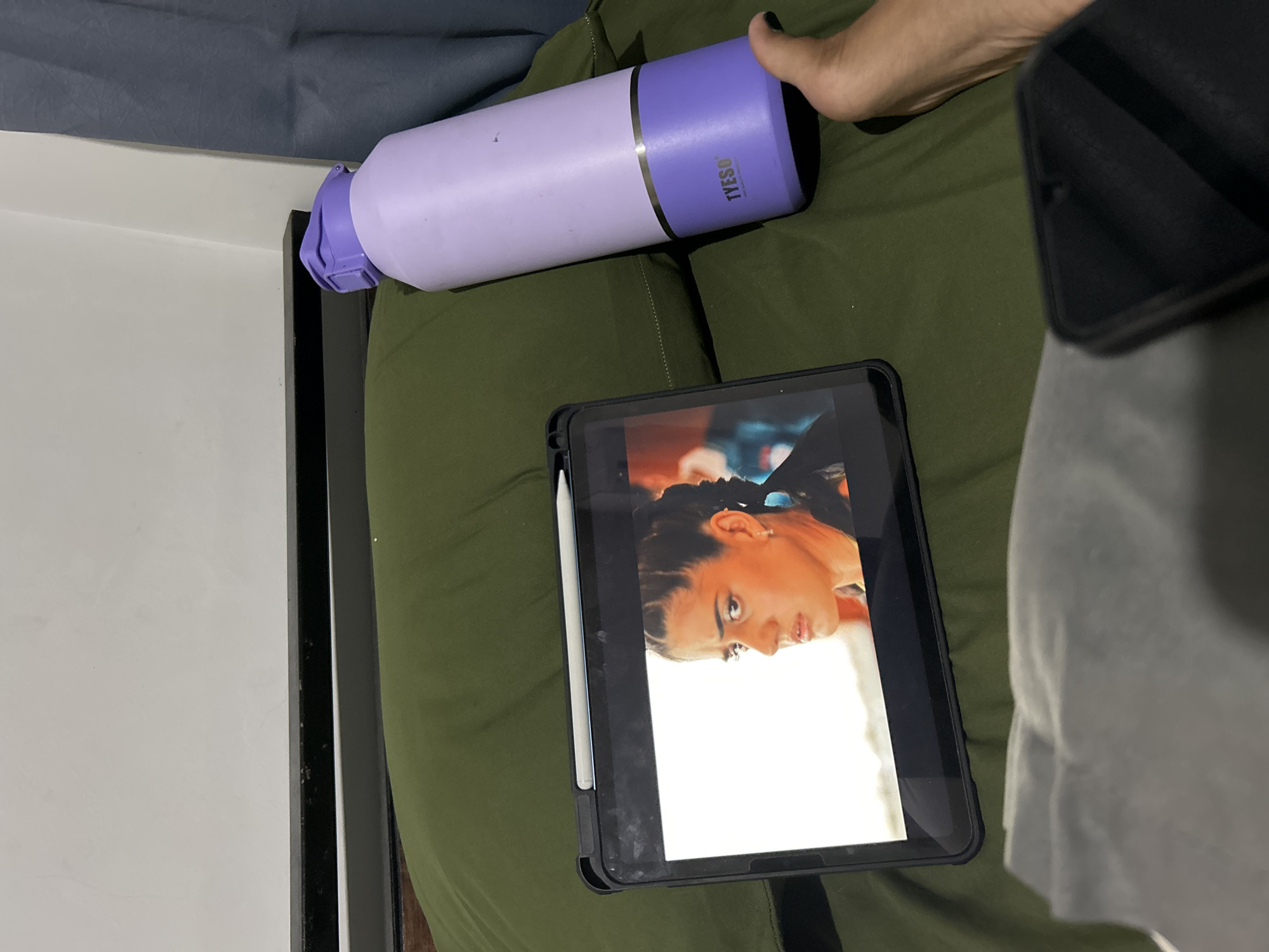 The image shows a person's hand holding a purple and light purple water bottle with the brand name "TYESO" visible on it. The water bottle is placed on a green, textured surface, possibly a blanket or clothing. In front of the water bottle, a tablet device is resting, displaying a close-up image of a woman's face. The woman has braided hair, is wearing earrings, and is looking towards the camera with a neutral expression. A white stylus is attached to the side of the tablet. To the right of the tablet, a black object, possibly a phone or another device, is partially visible. The background shows a dark gray fabric above a white surface and a black vertical border. The overall scene appears to be indoors, with natural lighting. The image composition is top-down, with the water bottle and tablet being the main subjects.