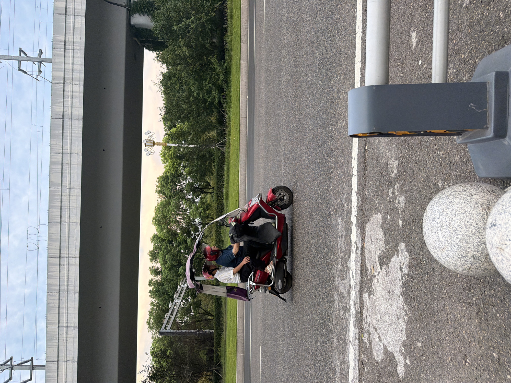 In Jiangjin, China, on a paved road under an elevated overpass, two individuals are riding a red three-wheeled electric scooter. The person in the rear, wearing a blue shirt and a red helmet, operates the vehicle. The passenger in front, wearing a white shirt and a pink helmet, is seated with their legs extended. The scooter has a canopy to shield them from the elements. The road is bordered by a grassy median with trees and vegetation behind it. Overhead, power lines and a concrete structure of the overpass are visible against a cloudy sky. In the foreground, two spherical concrete bollards are present.