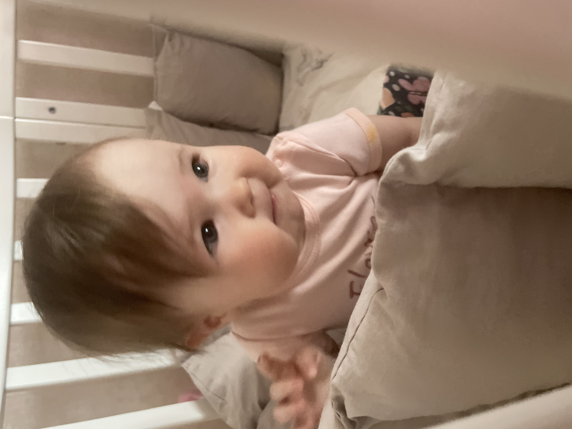 A baby lies in a crib. The baby has fair skin, brown hair, and blue eyes. The baby is wearing a pink t-shirt. A light-colored blanket partially covers the baby. Pillows are visible near the top of the crib. The crib has white slats. The location is indoors. There is no evidence suggesting this photo was taken in the city of Chaikovsky, Russia.
