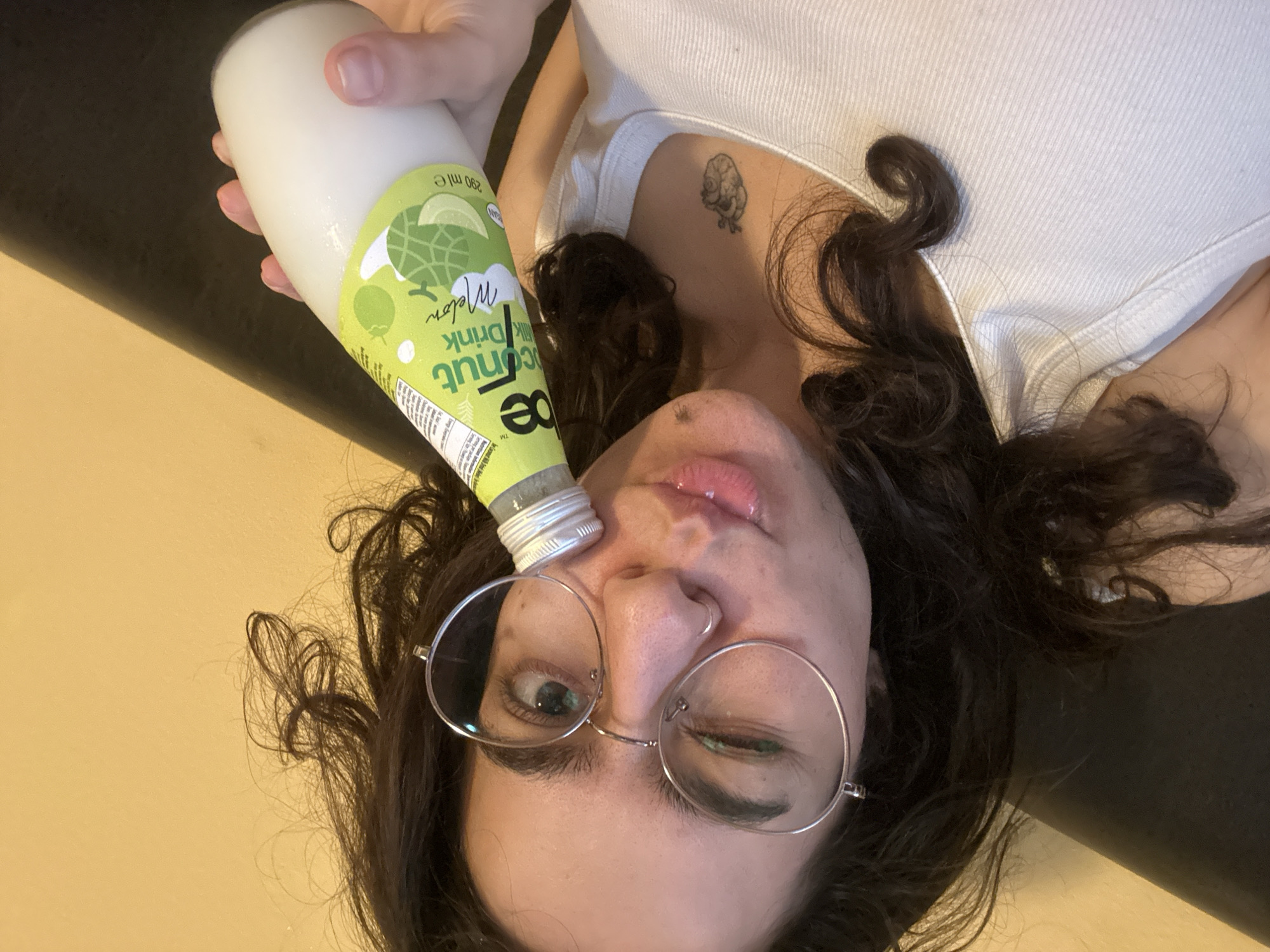 A person is holding a bottle of ae coconut drink. The person is lying down and looking at the camera. They are wearing round glasses. There is a tattoo on their chest. The background is a dark and light brown surface. There is no indication of the location being in Sofia, Bulgaria.