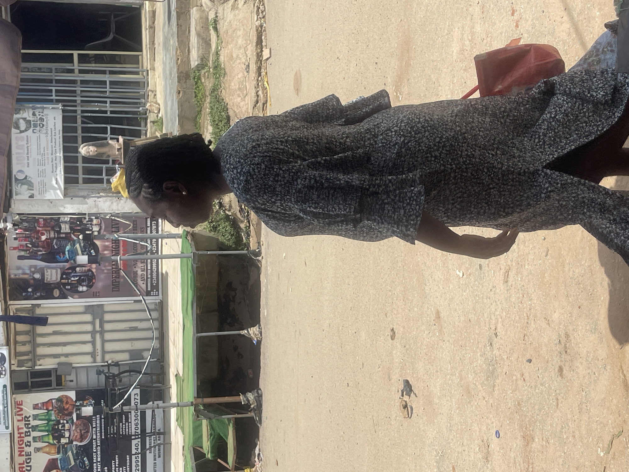 A woman in a gray patterned dress walks on a sunlit street in Abuja, Nigeria. She carries a red bag. Behind her are businesses, including "JOLA'S Hair and Beauty Saloon" and "IMPERIAL NIGHT LIVE LOUNGE & BAR." The buildings appear to be of concrete construction. There is scaffolding in front of the lounge and bar.