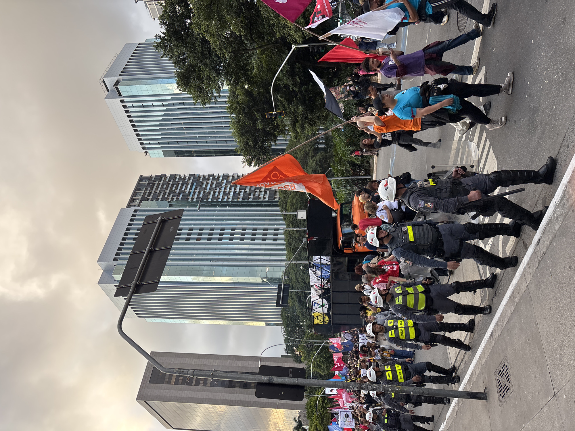 The image depicts a protest in São Paulo, Brazil, lined by police officers wearing helmets and protective gear. Protesters with flags are walking in the street. Behind them is a tall skyscraper and trees. The sky appears to be overcast.
