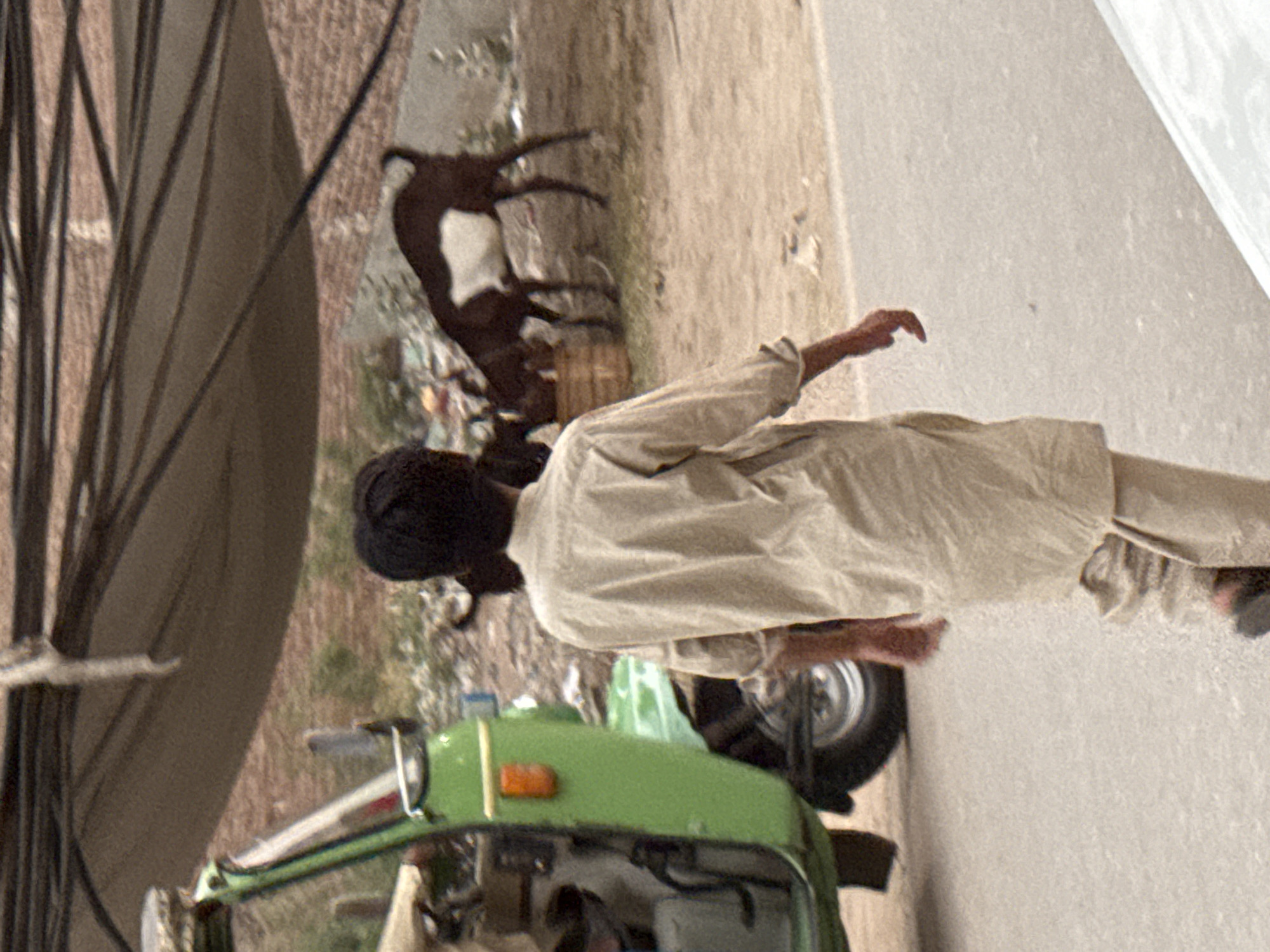 The image depicts an urban scene with a person dressed in a light-colored garment, possibly a traditional shirt, standing near what appears to be a green auto rickshaw or similar vehicle. The vehicle is in focus in the foreground and seems to be in operation. In the background, there are goats standing in a dusty area. The overall environment suggests a somewhat impoverished or rural-urban setting. A portion of a bicycle wheel is visible in the upper left corner, potentially indicating the presence of more vehicles or the nature of transportation in the area. The presence of trash on the ground near the goats contributes to the sense of environmental conditions. The image provides no definitive information about the city or country, so it is impossible to ascertain the location based on the information provided.