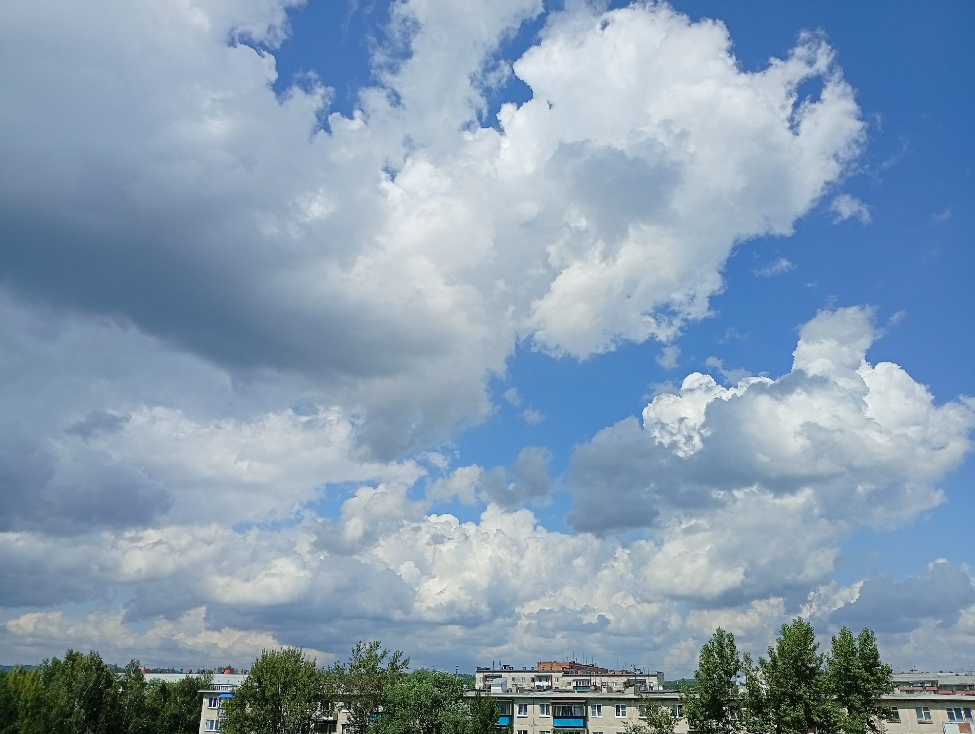 The image shows a view of the sky above Kyshtym, Russia. The sky is a mix of bright blue and large, puffy white and gray clouds. In the lower part of the image, buildings with several stories are visible, interspersed with green trees. The buildings appear to be residential apartment blocks. The overall scene suggests an urban environment with natural elements.