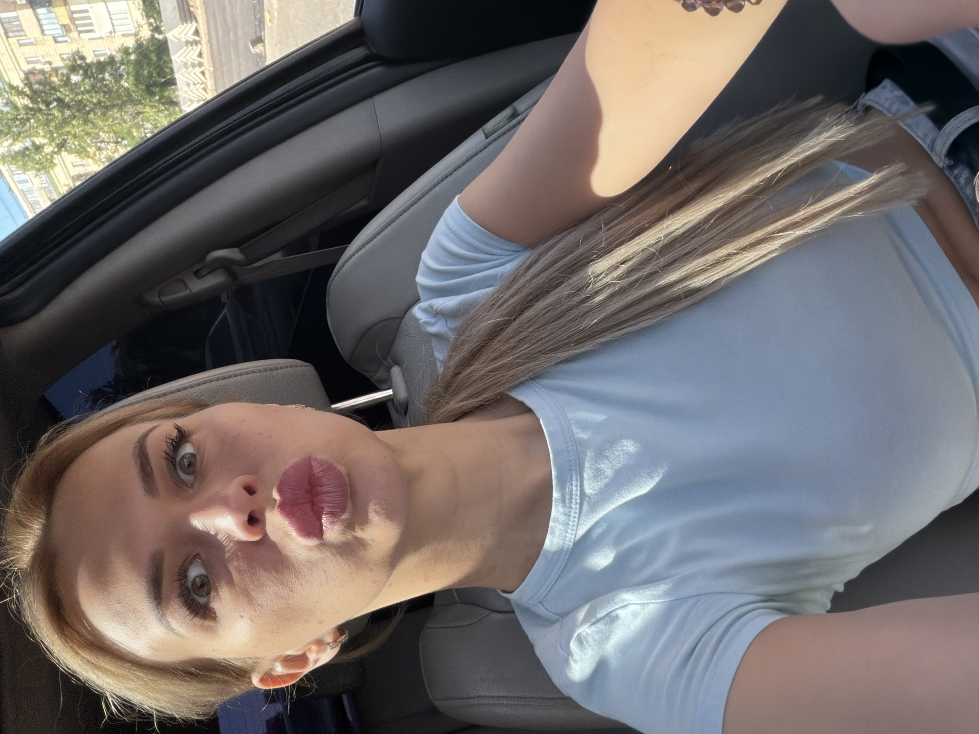 A young woman with blonde hair pulled back in a ponytail, wearing a light blue t-shirt, is captured in a selfie taken from inside a car. She is in the driver's seat, with the car's interior visible, including the seat and a portion of the dashboard. Her mouth is pursed, and her eyes are wide open, suggesting a playful or surprised expression. Sunlight streams into the car, illuminating her face and hair. Outside the car, through the window, a city street scene is visible, featuring buildings, trees, and construction materials, indicative of an urban environment in Kyiv, Ukraine.
