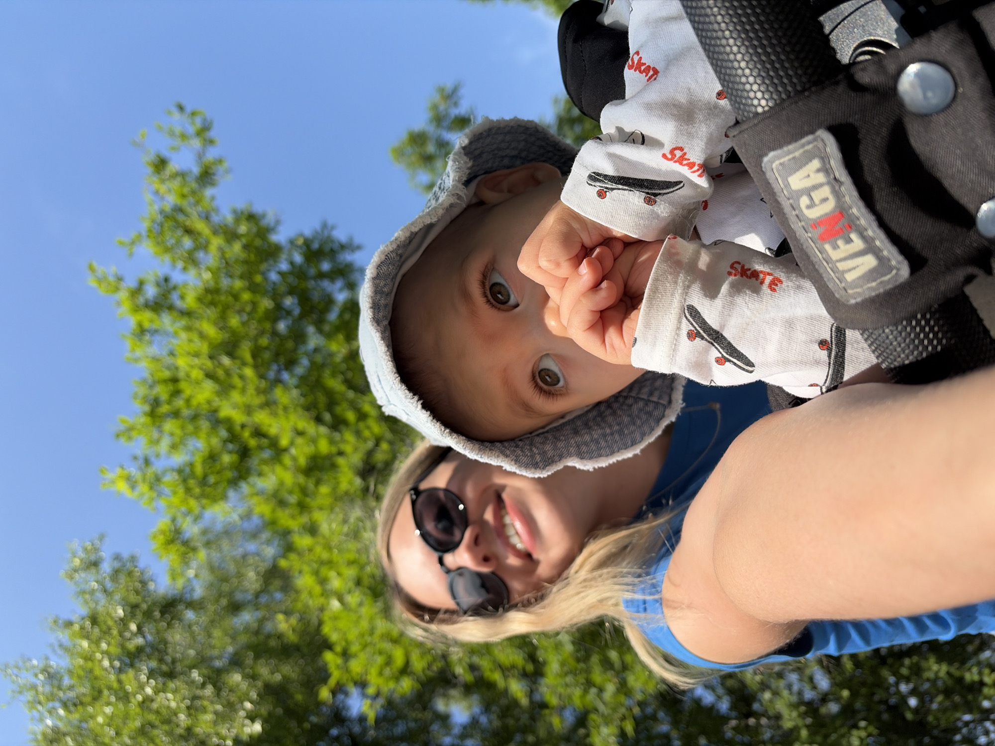 In a close-up, selfie-style outdoor shot, a young woman with blonde hair and sunglasses smiles broadly while holding a baby. The baby, wearing a light blue bucket hat and a white onesie with a skateboard print and "Skate" text, looks upward with its hands near its mouth. The scene appears to be in a park or natural setting, with lush green trees and a bright blue sky visible in the background. The woman is wearing a blue t-shirt, and part of a stroller handle with a "VENGA" logo is visible in the foreground. The image is taken from a low angle, with the sun shining brightly. There is no indication of the location being in Chaikovsky, Russia.