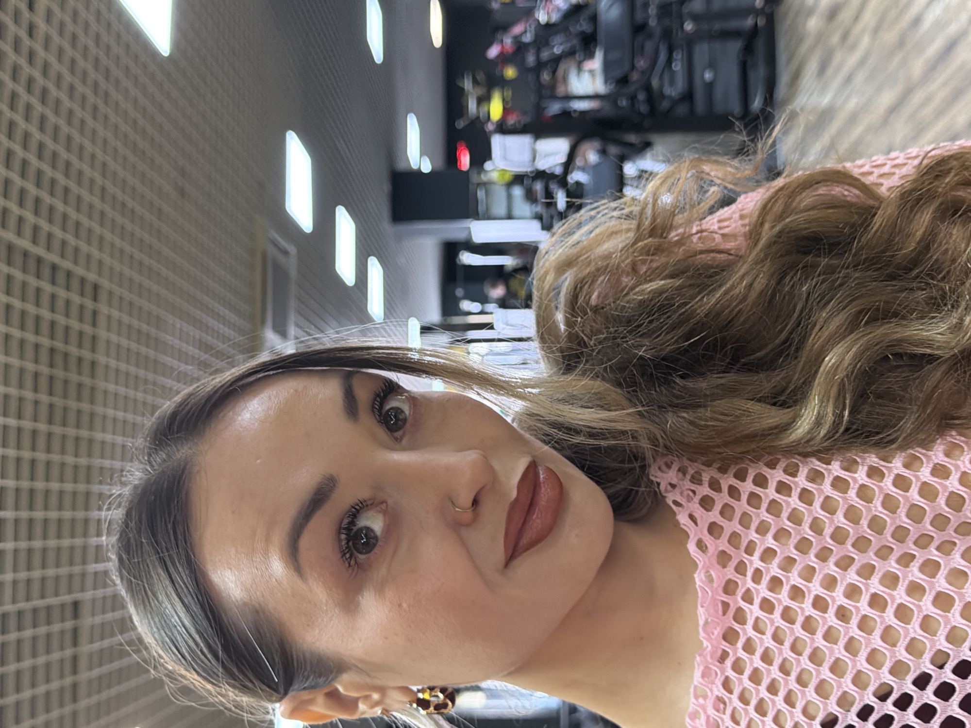 A portrait-style image, taken indoors in Krasnoyarsk, Russia, features a woman looking directly at the camera. She has long, wavy brown hair with lighter highlights, styled to one side, partially revealing a pink mesh or net-like top. Her face is well-lit, highlighting her makeup, which includes defined eyebrows, mascara on her eyelashes, a gold nose ring, and a matte, reddish-brown lipstick. She wears a patterned earring on her visible ear. Her expression is neutral and engaging.
The background shows an interior space with a distinctive ceiling composed of a light-colored grid pattern, possibly acoustic panels, interspersed with bright, rectangular overhead lights. Beyond the woman, the background is blurred, showing dark shapes that suggest gym equipment or salon furniture, hinting at a commercial or institutional setting. The image is taken at eye level, focusing on the woman's face and upper body. There is no visible text in the image.