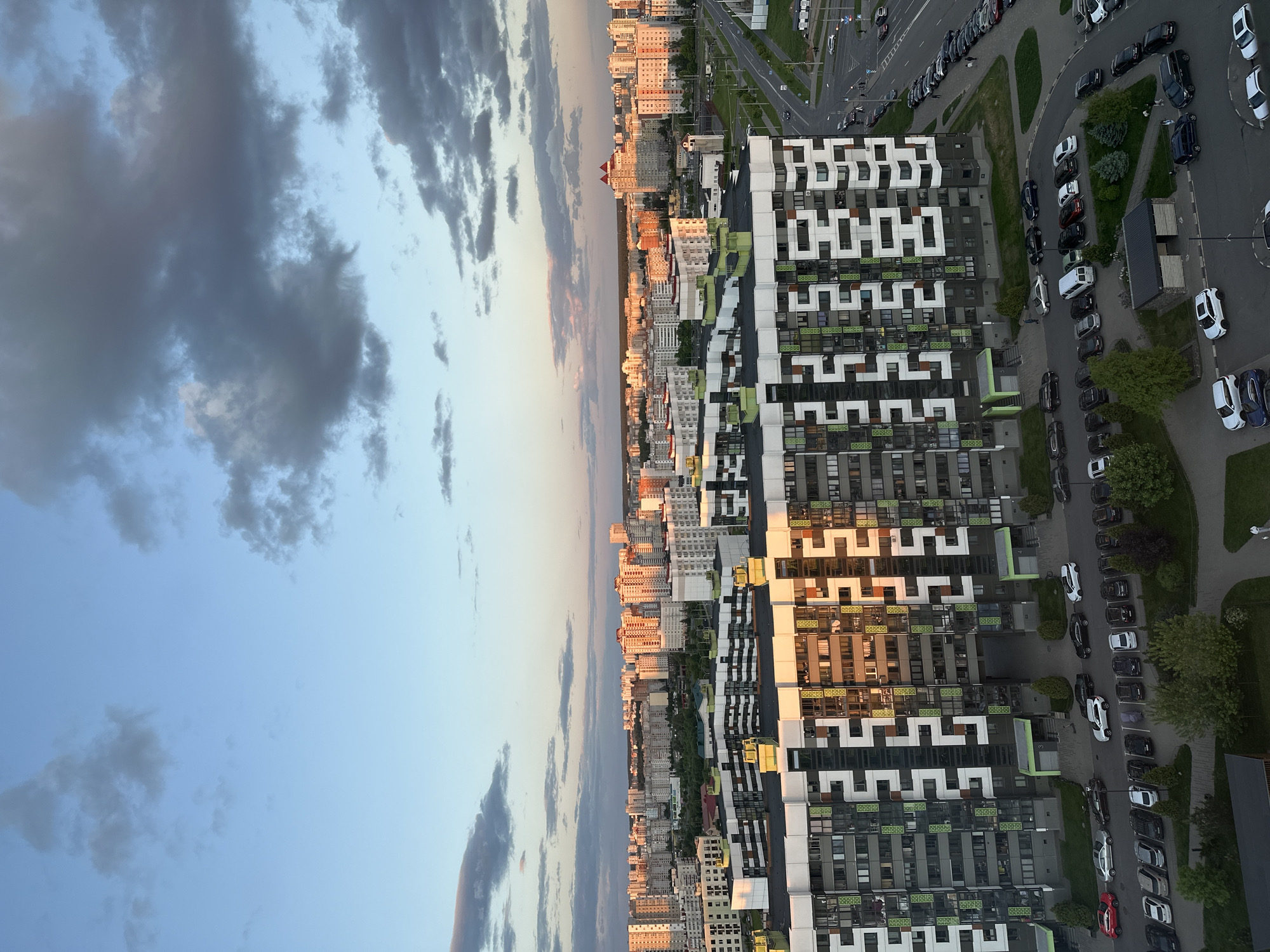 The image depicts an aerial view of a cityscape in Бараўлянскі сельскі Савет, Belarus. On the right side of the image, there are modern apartment buildings with a distinctive pattern of rectangular windows and green accents. Adjacent to the buildings, a road runs along the right side of the image, with numerous parked cars.
To the left of the modern apartment buildings, a more traditional city skyline stretches out, with buildings of varying heights and architectural styles. In the background, the sky is partly cloudy, with patches of blue sky visible between the clouds. The image is taken during the day, with soft, warm lighting on the buildings.