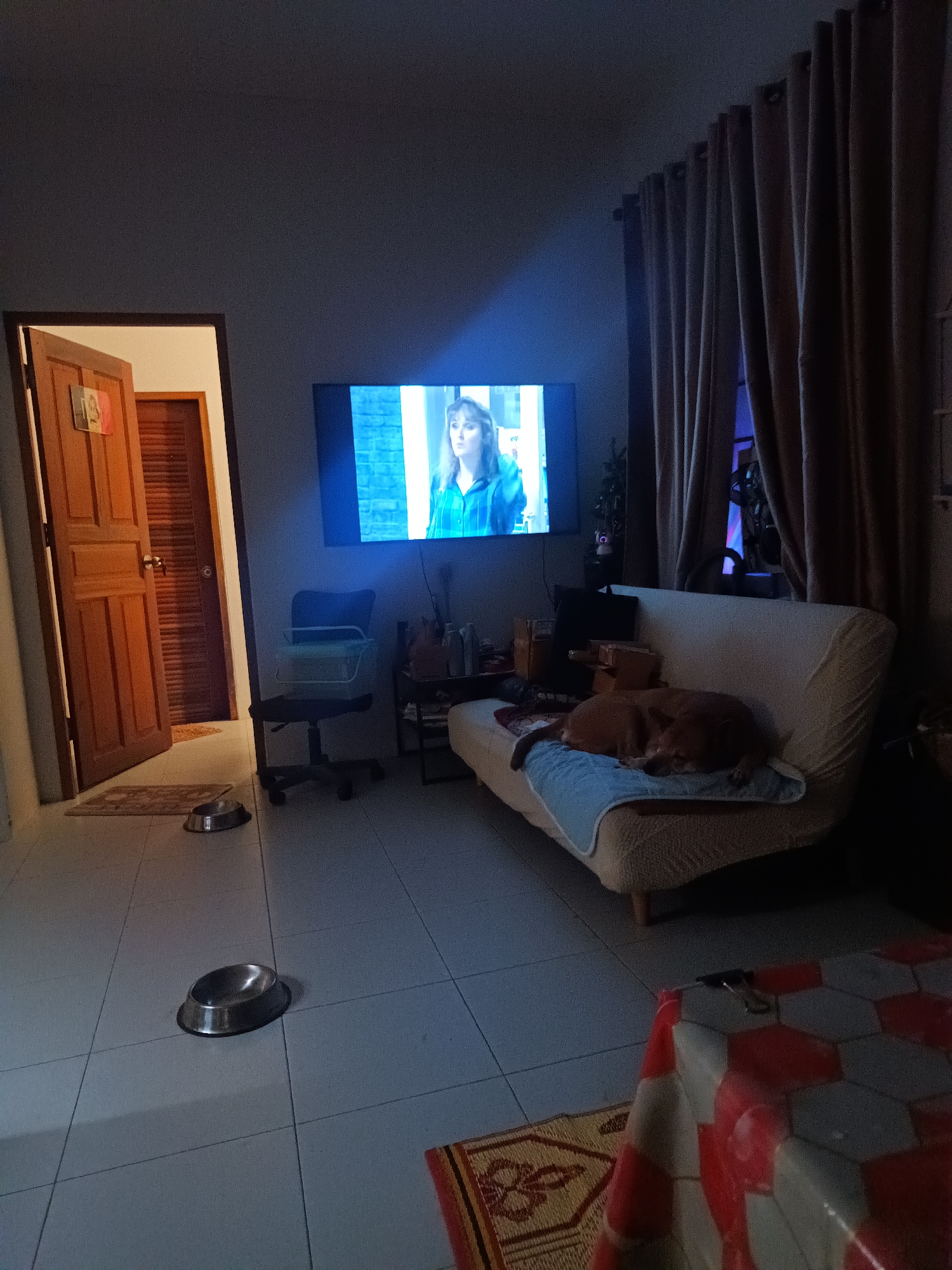 The image depicts the interior of a room in Moo 4, Thailand. The room is illuminated by a television screen showing a woman with blonde hair wearing a blue shirt. There is a brown dog asleep on a beige couch with a blue blanket. Two stainless steel dog bowls sit on the tiled floor. A desk chair is near the television. Curtains hang at a window. A partially opened wooden door leads to another room. A table with a red and white patterned cover and a patterned rug are in the foreground.