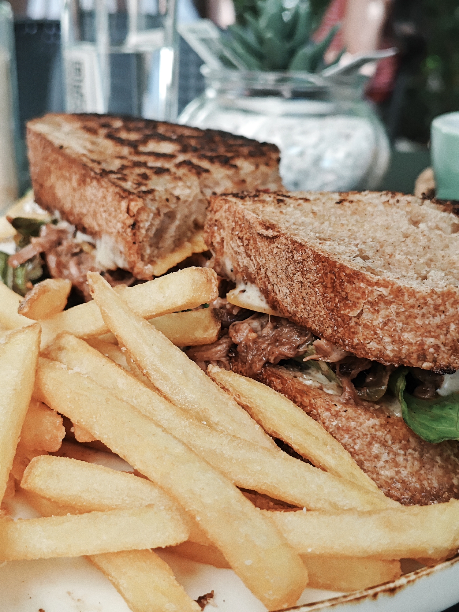 In an indoor setting with a shallow depth of field, a close-up shot features a plate of food. Dominating the foreground are numerous golden-brown french fries, some appearing lightly salted. Behind the fries, a sandwich is presented, sliced in half. The sandwich consists of thick, toasted bread filled with shredded meat, lettuce, and a creamy sauce. The background is softly blurred, showing a glass bottle, a glass jar with a succulent plant inside, and a teal-colored object, suggesting a restaurant or cafe environment. The lighting is natural and soft, creating a warm ambiance.