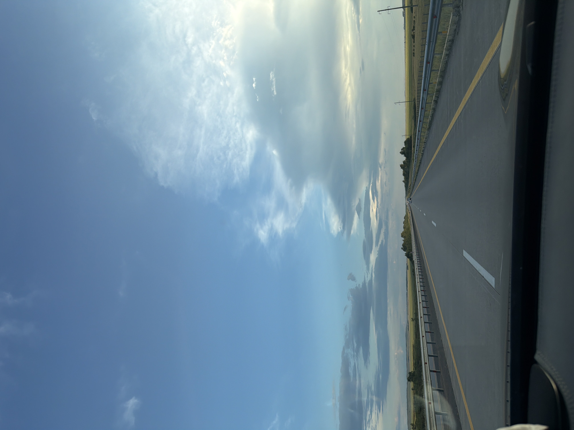 The image depicts a highway scene, likely taken from the perspective of a vehicle driving on the road. The highway stretches into the distance, with clear lane markings and a guardrail on the right side. The surrounding landscape appears to be flat and rural, with fields of grass or crops on either side of the road. Power lines are visible in the distance. The sky is partly cloudy, with a mix of blue sky and white and gray clouds. The lighting suggests it might be late afternoon or early evening. The location is specified as the city of Minovka, Kazakhstan. There are no people or specific subjects or objects that stand out besides the road and the landscape. The image is an objective record of a road trip scene.
