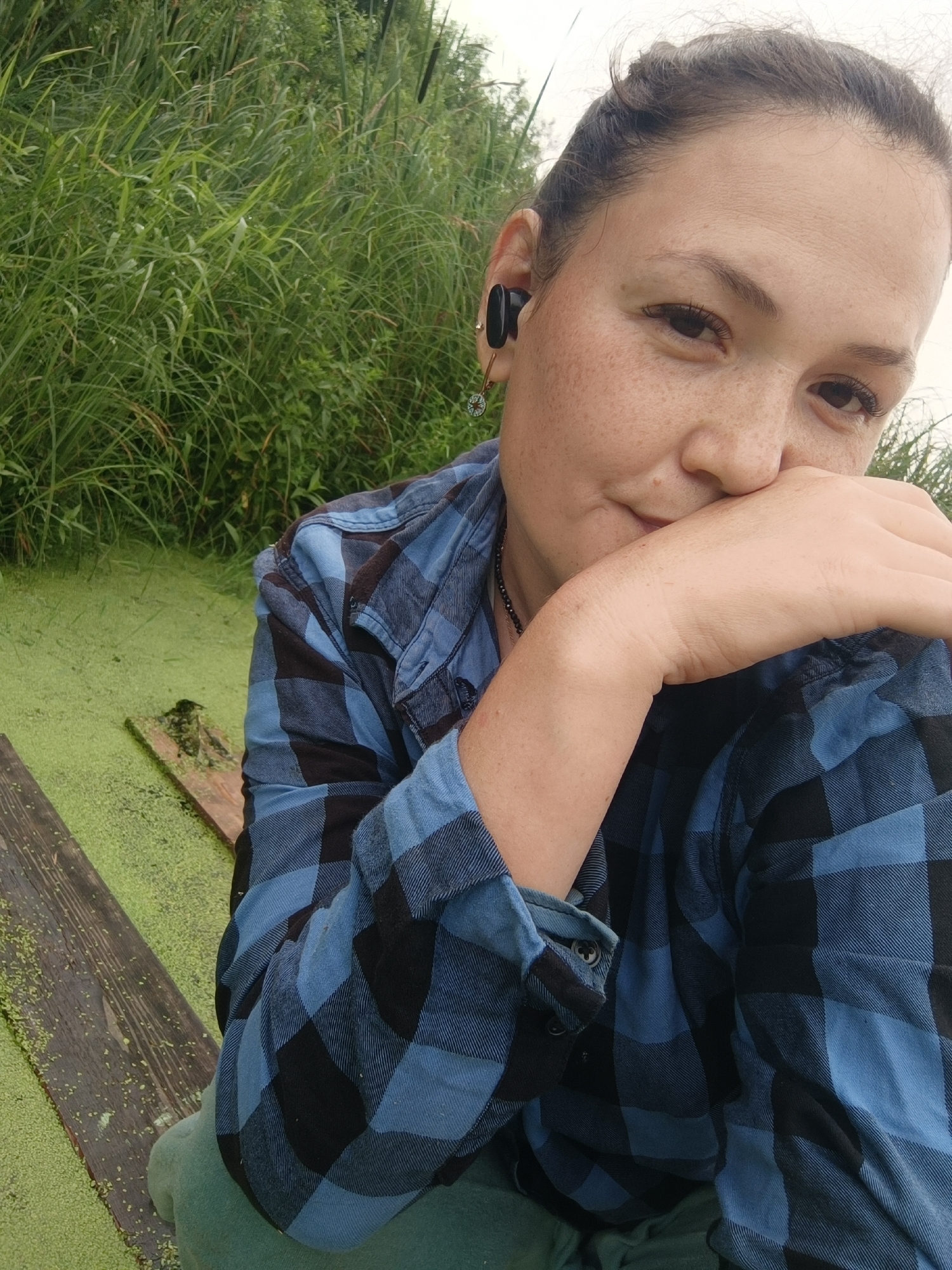 A close-up, waist-up, over-the-shoulder, outdoor, selfie style portrait of a woman. The woman has fair skin with freckles, brown hair styled in a braid, and is wearing a blue and black plaid shirt. She is looking at the camera with a slight smile and her hand covering her mouth. She is wearing a black earbud in her left ear. The background is filled with tall green grass and a body of water covered in green duckweed. The setting appears to be a rural or natural environment, possibly a pond or marsh, on the outskirts of the city of Karsha, Russia. There is a weathered wooden plank in the water to the left of the woman.