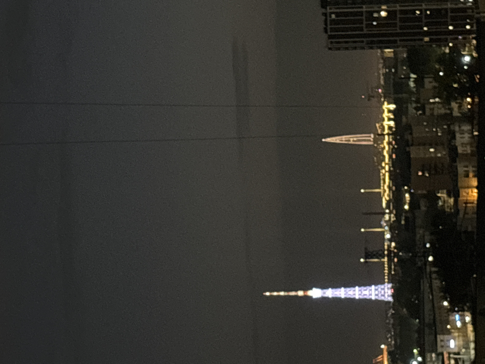 Captured at night in the city of Tokyo, Japan, the image shows a skyline dominated by the Tokyo Skytree, illuminated in purple and white lights. In the distance is Tokyo Tower, emitting a softer light. Buildings, trees, and streetlights are also visible. Overhead are thin black lines, likely electrical wires.
