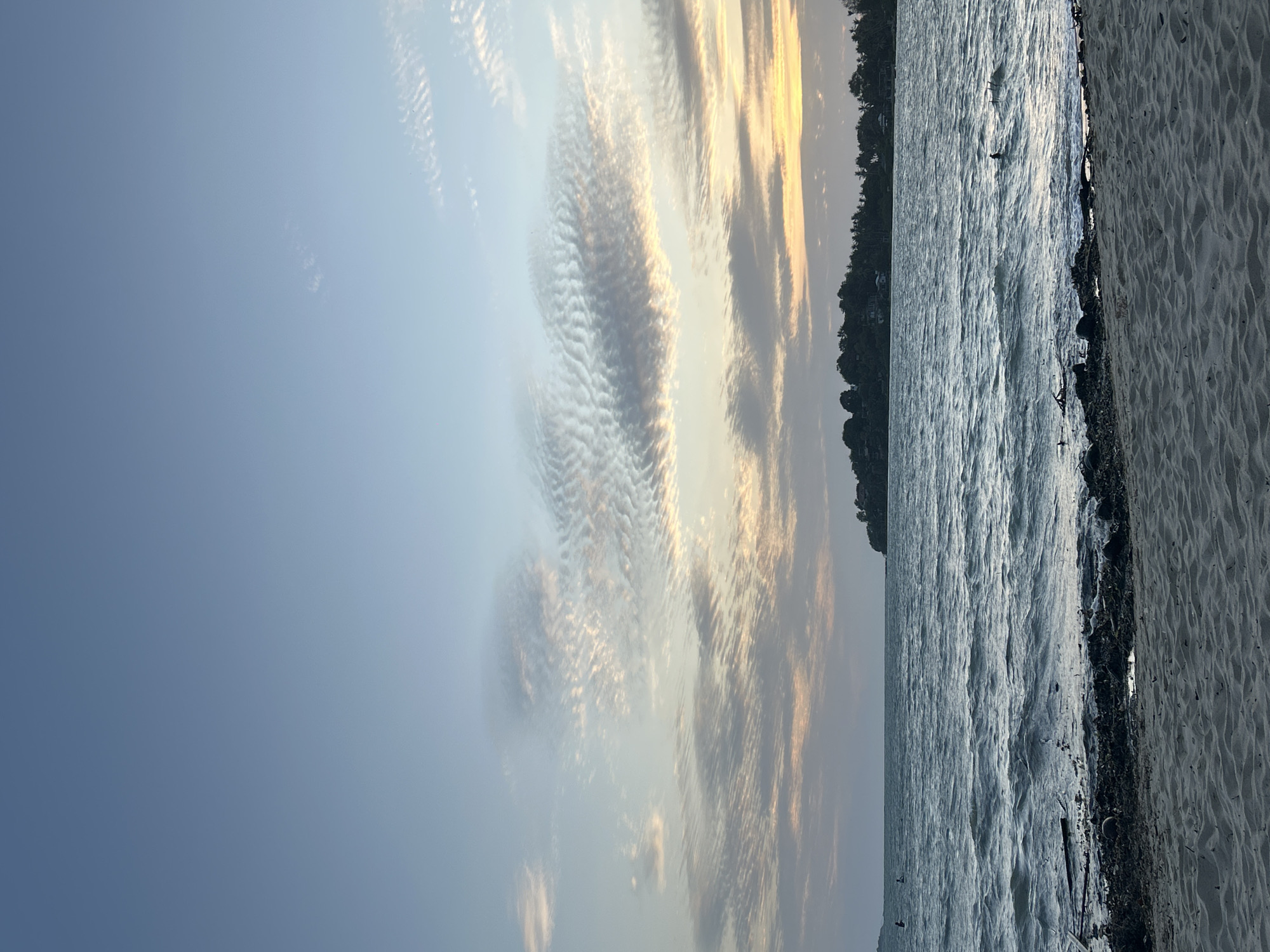 A tranquil beach scene unfolds with a wide expanse of light blue sky, tinged with pastel hues of orange and yellow from the setting sun. Wispy, rippled clouds drift across the horizon, creating a serene and atmospheric backdrop. Below, the deep blue water of the Gulf of Thailand gently laps at the sandy shore. The water is a little choppy, with small waves creating a textured surface. Debris, including driftwood and coconut husks, is scattered along the waterline, hinting at the natural ebb and flow of the tide. In the distance, a lush green coastline with scattered houses and palm trees forms a picturesque silhouette against the sky. The sandy foreground is textured with small ripples, suggesting the presence of wind. The scene captures a quiet moment at dusk, likely on a beach in the Ko Pha-ngan District of Thailand, a popular destination known for its stunning natural beauty and relaxed atmosphere.