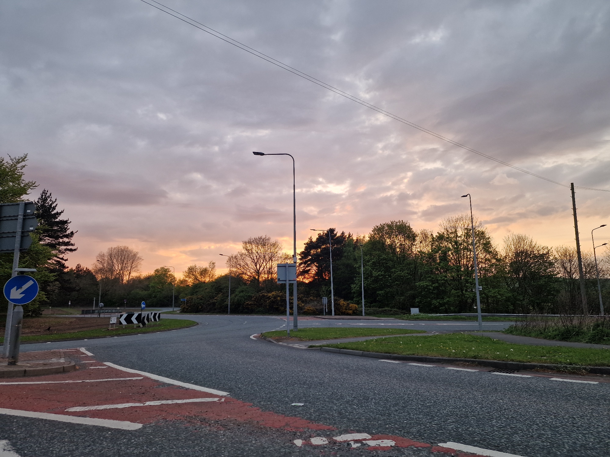 The image shows a road junction in Pity Me, United Kingdom. The road surface is asphalt, marked with red and white pedestrian crossing lines. A blue sign with a white arrow indicating a left turn is visible on a roadside post. A series of black and white chevron signs mark the bend in the road. There are trees and vegetation lining the roadside. Streetlights are present. The sky is overcast with a sunset glow. No people are visible.