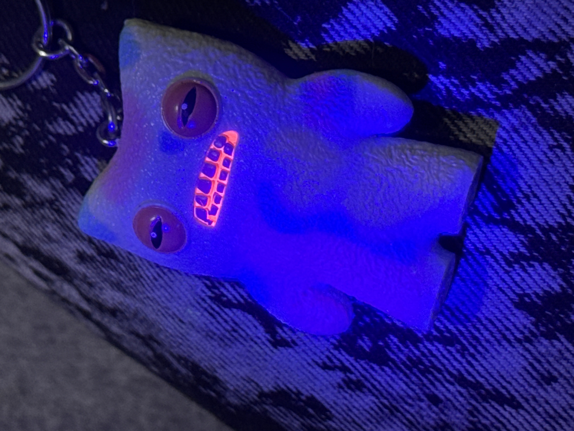 The image shows a blue, creature-shaped keychain with bulging eyes and bright orange teeth. The keychain is lying on a textured surface, likely denim. A chain is visible on the left side of the frame, attached to the keychain. The subject is illuminated by a blue or ultraviolet light. The environment suggests an indoor setting. There is no information to suggest that the image was taken in the city of Sofia, Bulgaria.