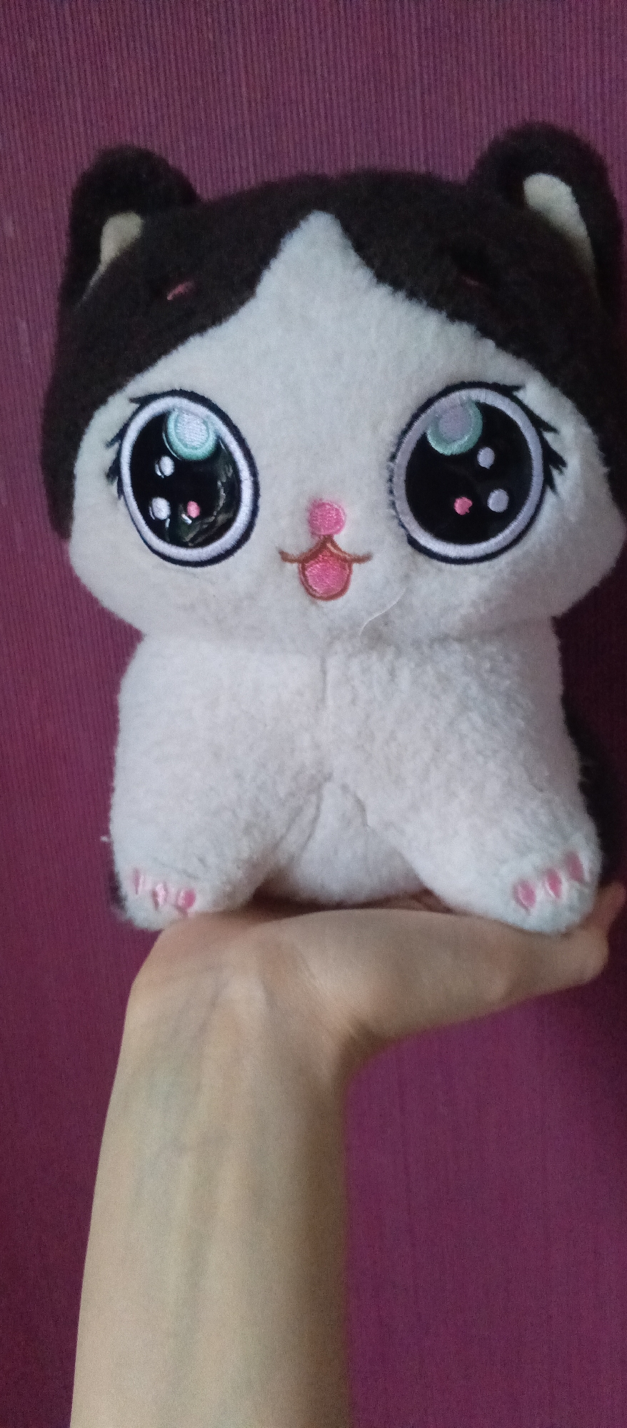 A person's hand is supporting a stuffed cat toy. The cat toy is primarily white with brown fur on its head and ears. It has large, round eyes with black irises, white highlights, and colorful iridescence that reflects light. The cat has a small pink nose and a stitched mouth that resembles a tongue. Its front paws are visible, each with three small pink toe beans. The toy is positioned on the palm of the person's hand, which is light-skinned. The background is a solid, textured purple surface, possibly a wall or fabric. The image appears to be taken indoors in natural or artificial light. No text is visible in the media file.
