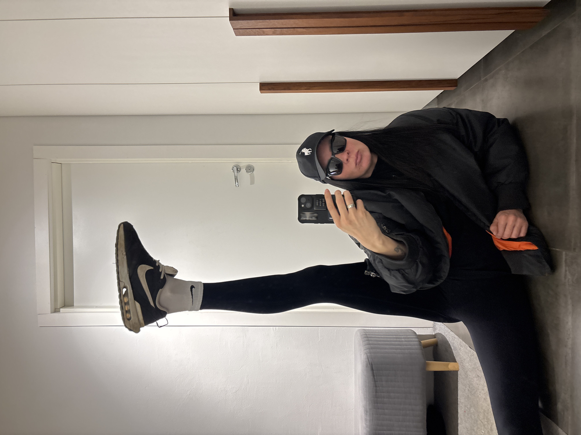 A woman in a black jacket, black pants, and black sneakers is taking a selfie in front of a mirror. She is wearing a black baseball cap with a white logo and black sunglasses. Her left leg is extended and resting on the mirror's edge, near the door. A small grey stool is visible in the foreground. The location is indoors, likely an apartment or house with white walls, a white door, and grey tiled flooring. There are wooden decorative elements on the ceiling or upper wall. Based on the query prompt, the implied location is Chaikovsky, Russia, although there is no visual confirmation of this within the image.