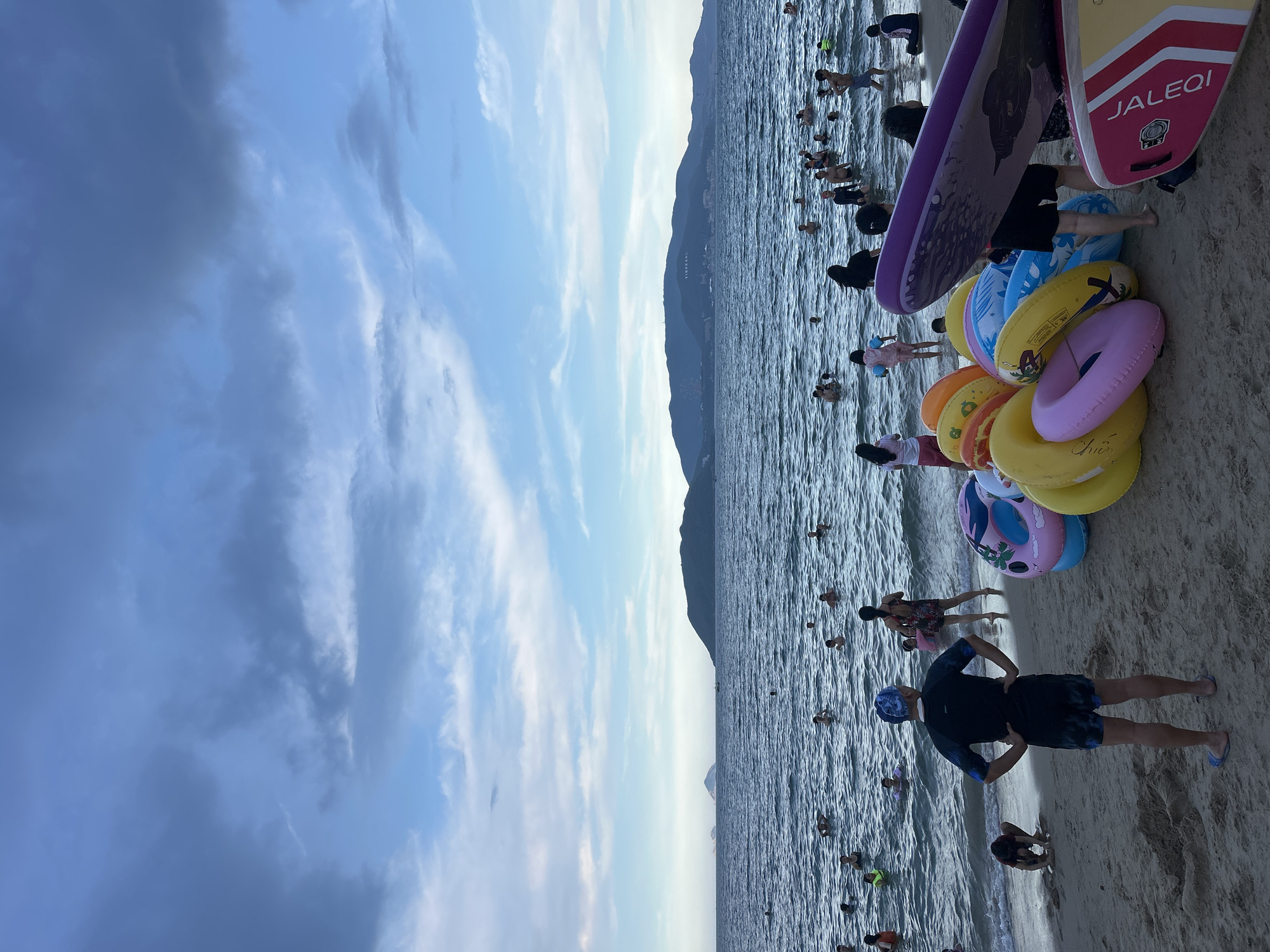 The image shows a crowded beach in Nha Trang, Vietnam. The sky is cloudy with patches of blue. The ocean is choppy and filled with people swimming and wading. On the beach, there are several inflatable tubes and two paddleboards piled up. A man in a black rashguard and patterned swim trunks stands facing away from the camera, hands on his hips. Another person is crouching on the sand. In the background, a coastline with mountains is visible.