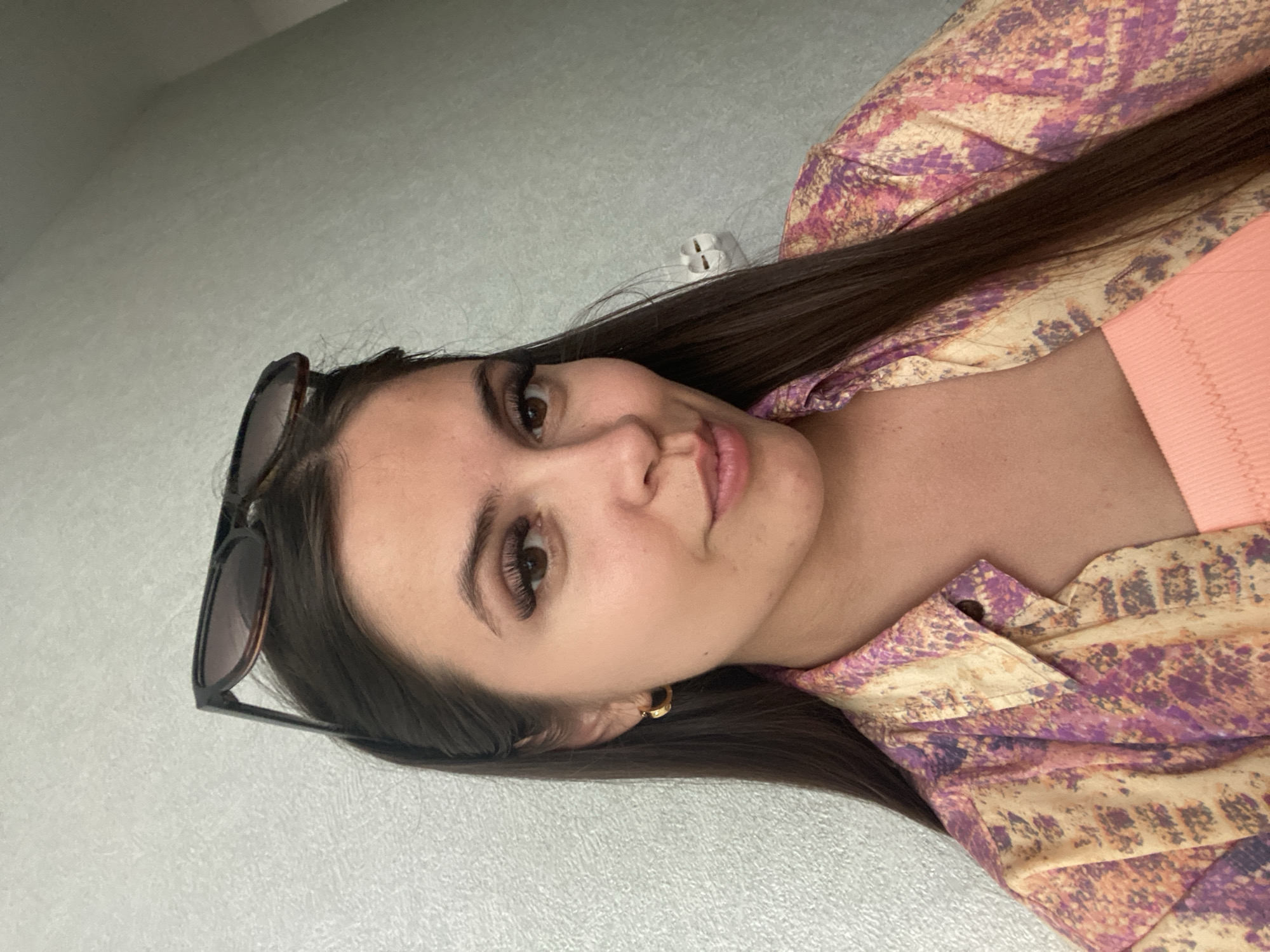A young woman with long, dark hair is pictured from the chest up. She is wearing a patterned button-down shirt over a coral-colored top, and sunglasses are perched on top of her head. A gold hoop earring is visible in her left ear. She is looking towards the camera with a slight smile. The background is a textured, light-colored wall with a white electrical outlet. The image appears to be taken indoors, likely in a residential setting, with no specific identifiable landmarks of Chaikovsky, Russia visible.