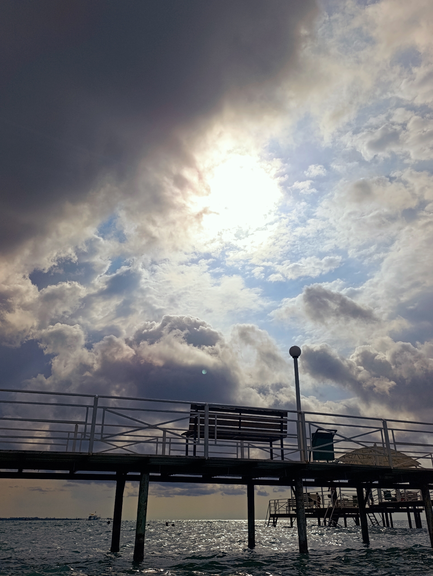 This is an outdoor, low-angle, eye-level, full shot of a pier extending over the water in Sary-Oy, Kyrgyzstan, under a dramatic sky. The sky is filled with dark, heavy clouds partially obscuring a bright sun, creating strong contrast and shafts of light. The water is choppy with sunlight glinting off its surface.
The pier is constructed of dark wood and supported by visible pilings. A white metal railing runs along the edge of the pier. On the pier, there is a wooden bench. Further down the pier, there appears to be a trash receptacle and a partially visible straw umbrella. There are a few individuals on the lower section of the pier, near the water's edge, with one person descending a ladder. In the distance, a small boat is visible on the water.