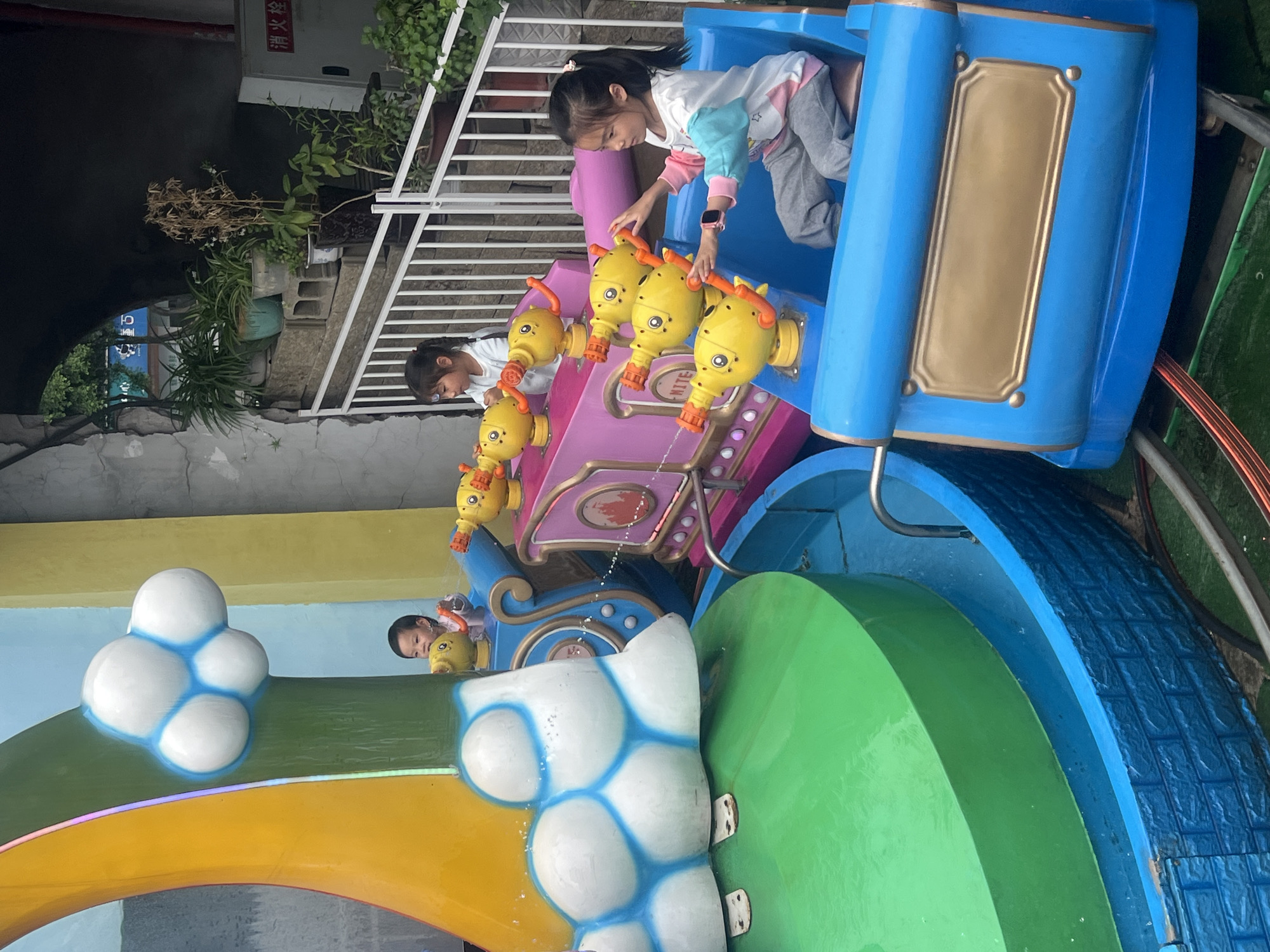 Two children are playing with water jets on a ride at the Nan'an District, China park. The ride features colorful train cars, water-squirting animal figures, and decorative elements like clouds and a rainbow.