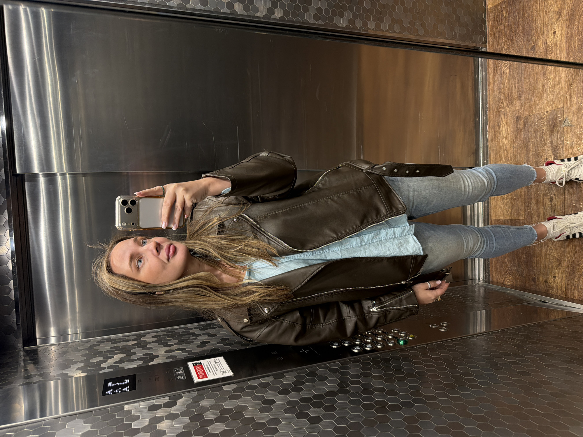 A woman is taking a selfie in an elevator. She is wearing a brown leather jacket, light blue shirt, jeans, and white sneakers with red stripes. The elevator has a patterned floor, stainless steel walls, and buttons. It is not possible to confirm the specific location in Moscow, Russia, without additional information.