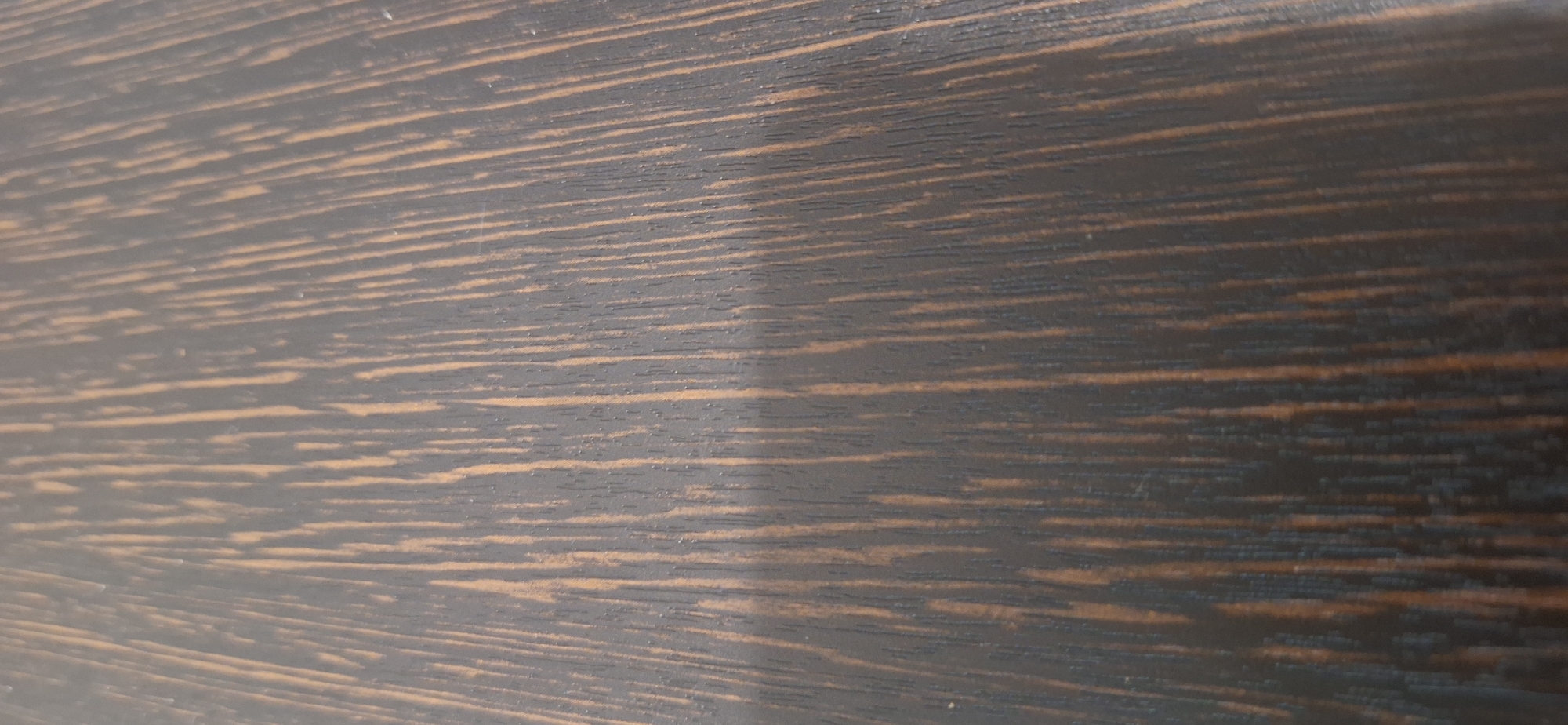 The image shows a close-up, angled view of a dark wood surface with a prominent grain pattern. The grain consists of numerous vertical, parallel lines of lighter brown alternating with wider, darker brown or black areas. The surface appears to be a tabletop or a similar flat wooden object. There are no people, buildings, or any other environmental context visible. The lighting suggests it is indoors or under artificial light, casting subtle shadows that emphasize the texture of the wood. There is no information in the image that can be used to determine its location in Bila Tserkva, Ukraine.