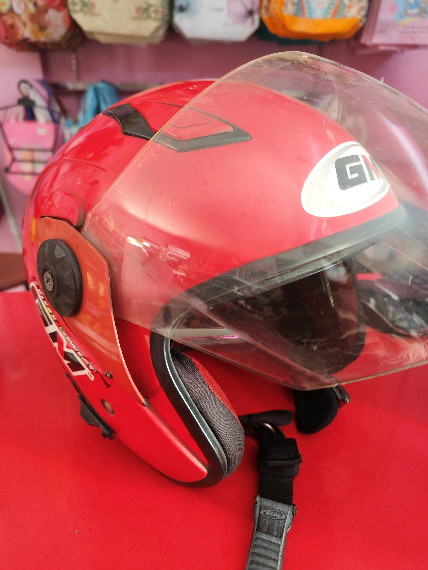 A close-up, eye-level, vertical shot shows a red helmet with a clear visor resting on a red surface, likely a table or floor. The helmet is angled, revealing its side and front. A black strap with a silver buckle is visible, extending from the helmet's chin strap. The background is blurred, showing colorful items that could be bags or merchandise. The lighting is even, casting soft shadows. The image is sharp and detailed, highlighting the texture of the helmet and strap. No people are present in the image. The image was taken in Sragen, Indonesia.