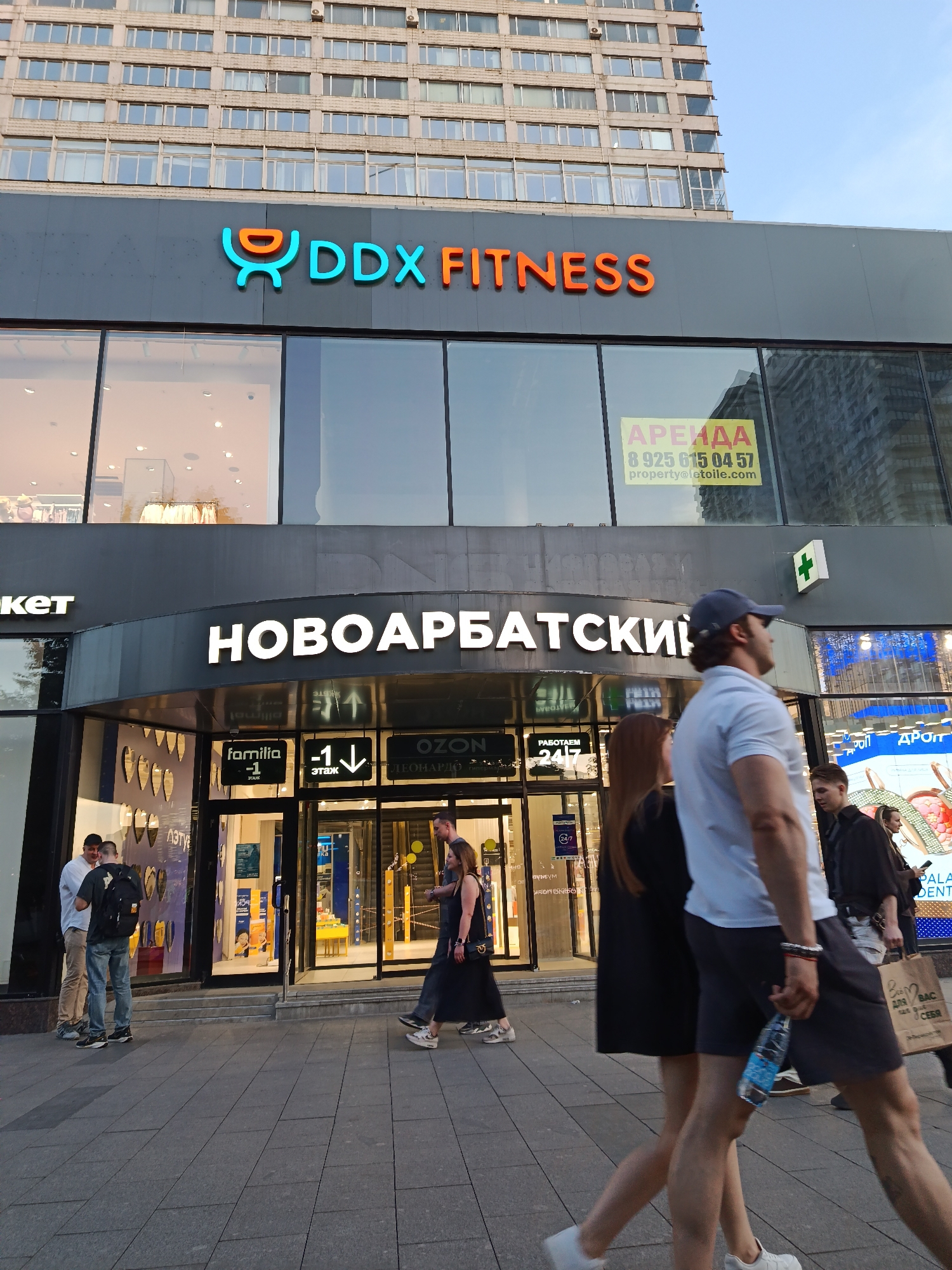 This image captures a street-level view of a multi-story commercial building on Novy Arbat Street in Moscow, Russia, featuring pedestrians on the sidewalk.
The upper portion of the building consists of numerous floors with a grid-like arrangement of windows, presenting a light-colored, possibly beige or grey, facade. Below this, the building transitions to a dark grey commercial facade with large glass storefronts. Prominently displayed is the "DDX FITNESS" logo in blue and orange lettering, with a stylized 'DDX' symbol. Beneath the fitness sign, a large white illuminated sign reads "НОВОАРБАТСКИЙ" (Novy Arbat).
Within one of the storefront windows, a yellow "АРЕНДА" (RENT) sign with a phone number and email "property@letoile.com" is visible. Near the building's recessed entrance, additional signs indicate various businesses: "familia -1 ЭТАЖ" (Familia -1 Floor), "OZON," and "ЛЕОНАРДО" (Leonardo). A green cross symbol, typically denoting a pharmacy, is affixed to the building's exterior on the right side. Another sign states "РАБОТАЕМ 24/7" (Working 24/7).
Approximately eight to ten individuals are present. In the immediate foreground, a man wearing a white short-sleeved polo shirt, dark shorts, and a blue baseball cap walks from right to left, carrying a clear plastic water bottle in his left hand. To his left, a woman in a black dress walks in the opposite direction. Further back on the right, another man with a dark shirt holds a brown paper bag. Near the building's entrance on the left, two men are standing; one wears a light shirt and beige trousers, the other a dark shirt, jeans, and a black backpack. Other indistinct figures are visible through the glass doors and windows of the commercial establishments.
The sidewalk is paved with grey rectangular tiles. The lighting suggests daylight conditions, casting soft shadows. The sky, visible in the upper right, is a clear light blue.