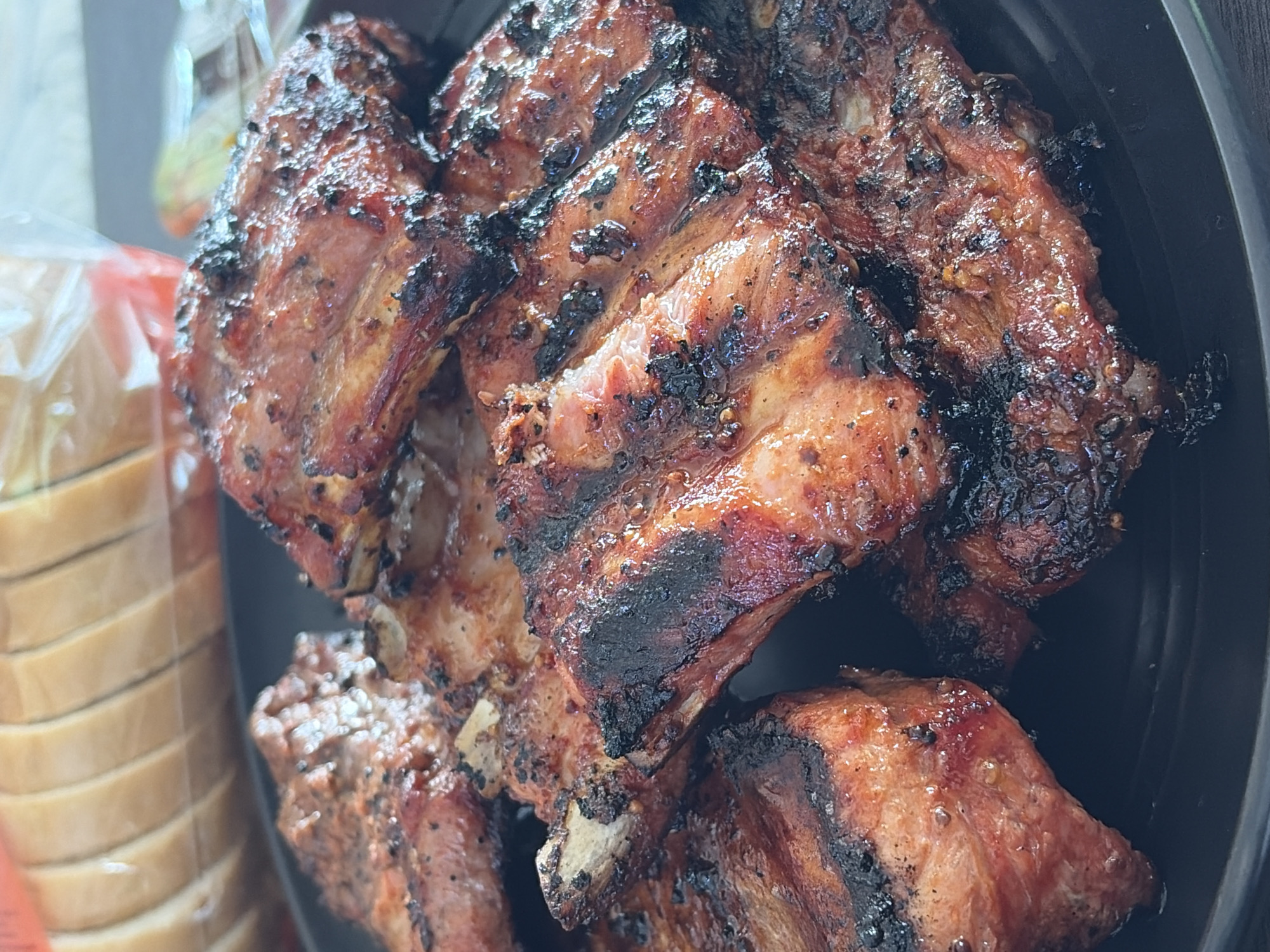 A close-up, overhead shot shows a pile of grilled pork ribs on a black plate. The ribs are coated in a dark, glistening sauce and have char marks from the grill. Some of the bones are visible. In the background, out of focus, is a package of sliced white bread. The image does not provide any information about the city of Vidogoshchi, Russia, or any people present.