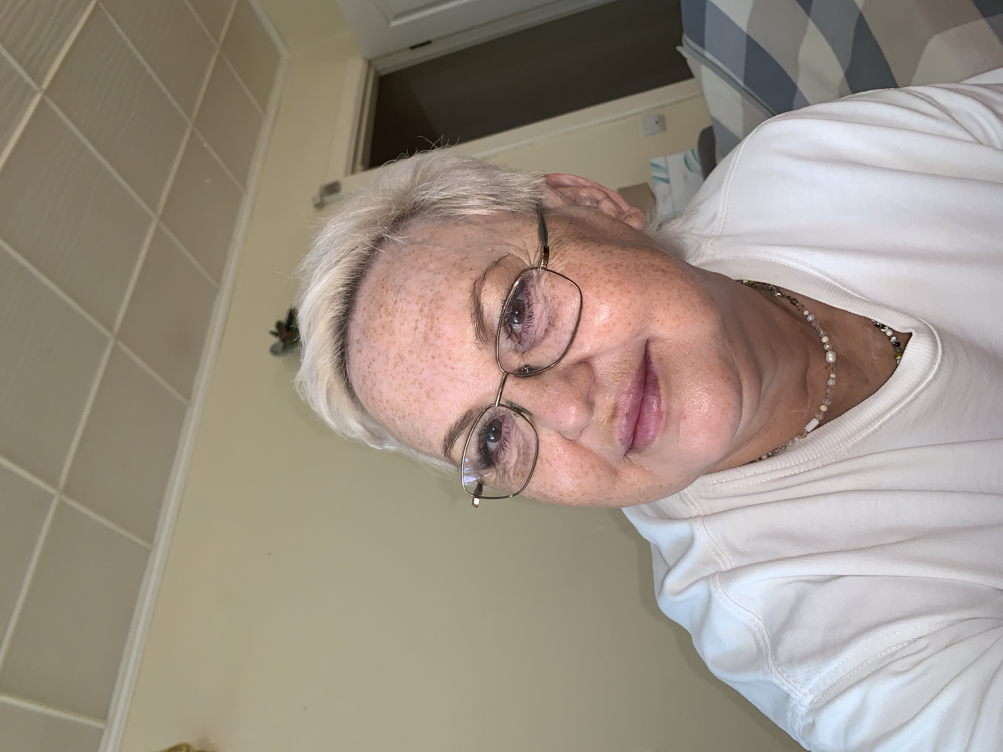 A close-up indoor shot features an older woman with white hair and freckled skin. She is wearing glasses and a white shirt, and a beaded necklace is visible around her neck. The background includes a tiled wall on the left, a door or window frame above her head, and a blue and white patterned object (possibly bedding) to the right. The image provides no information to suggest that this image was taken in the city of Chaikovsky, Russia.