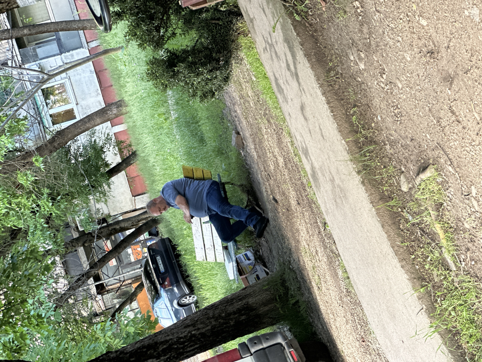 The image shows a scene in Lovech, Bulgaria. A man is sitting on a park bench next to a parked dark-colored car, with visible Bulgarian license plates. The setting includes trees, a grassy area, a sidewalk, and a glimpse of buildings. The man appears to be resting or looking at something in his hands. Several boxes are placed under the bench.