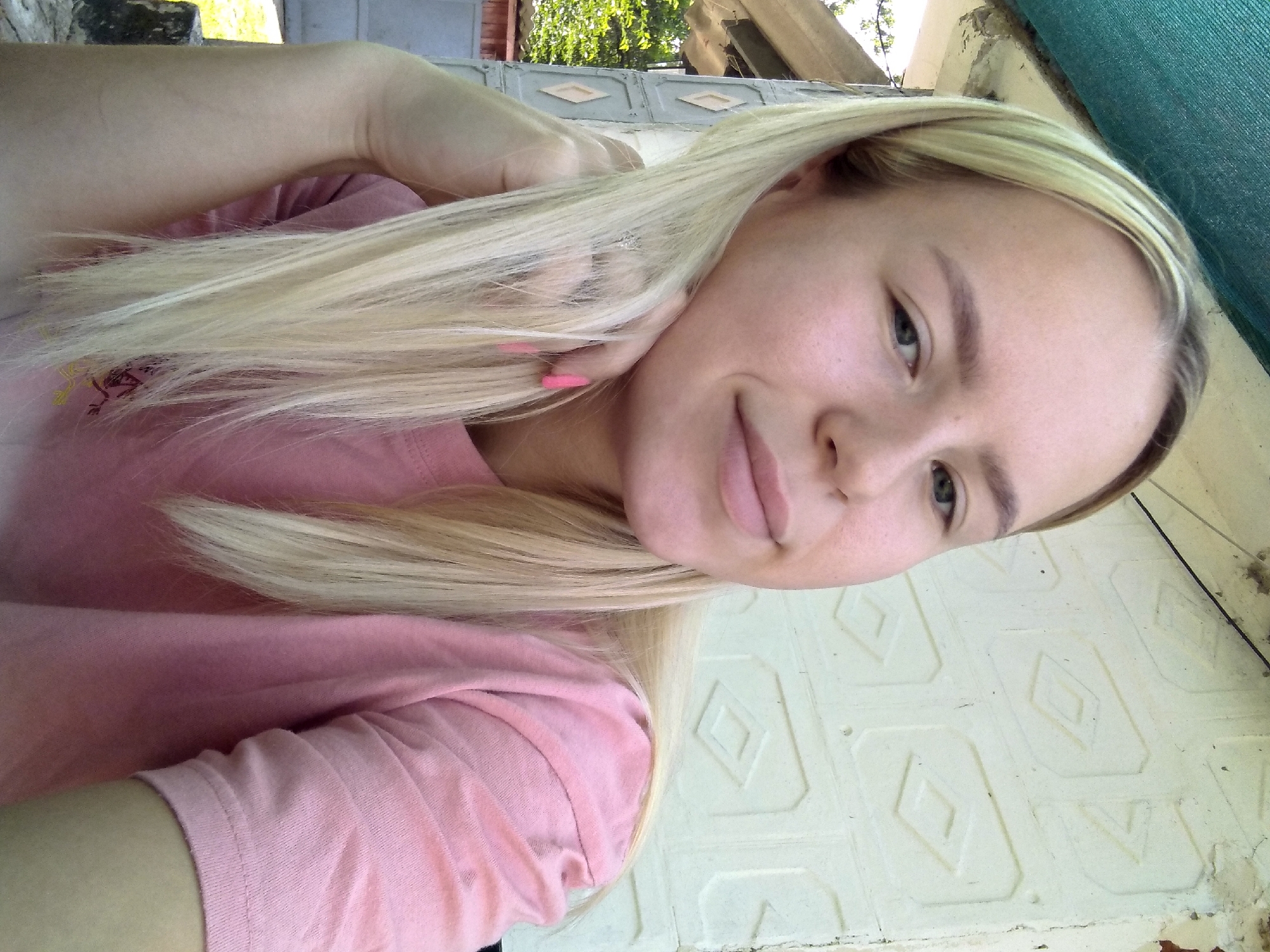 The image shows a young woman with blonde hair, wearing a pink t-shirt. She is posing for a selfie, resting her head on her hand. The background appears to be an outdoor setting with a textured wall and some greenery visible. The location is identified as Radisne, Ukraine.