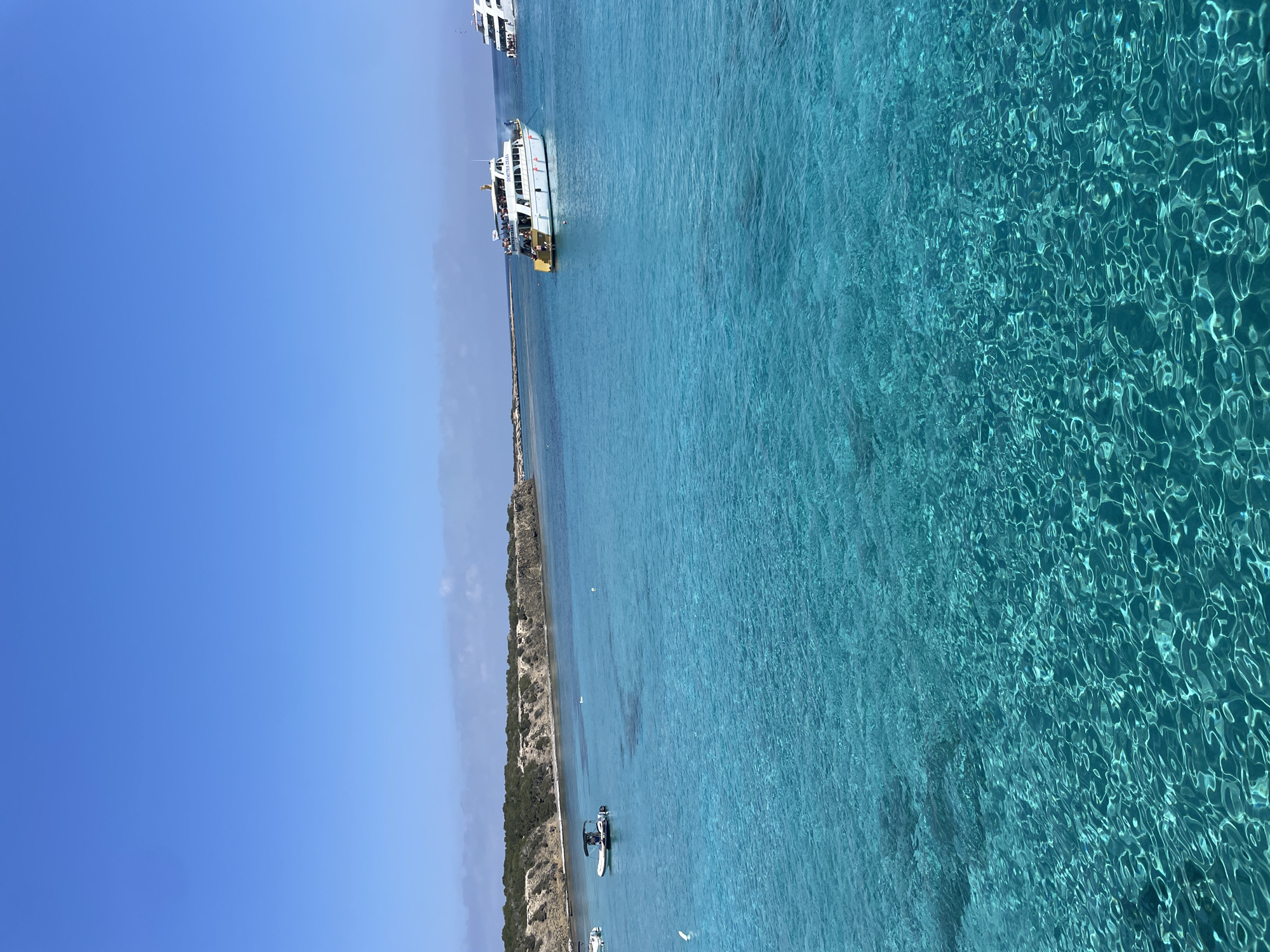 This is a vertical, eye-level, medium-shot photograph of the clear, turquoise waters of the Mediterranean Sea near Neo Chorio, Cyprus. The scene is dominated by the vibrant blue water, which is so transparent that the sandy seabed is visible beneath the surface, with ripples and light patterns dancing on the water. In the middle ground, a white tour boat named "DEMETRIS CHARA" is visible, carrying numerous passengers on its deck. To the left of this boat, a smaller inflatable dinghy with a canopy and an individual steering it is anchored. In the background, a rocky, tree-covered coastline stretches across the horizon. The sky above is a clear, bright blue, with a few wispy clouds near the horizon.