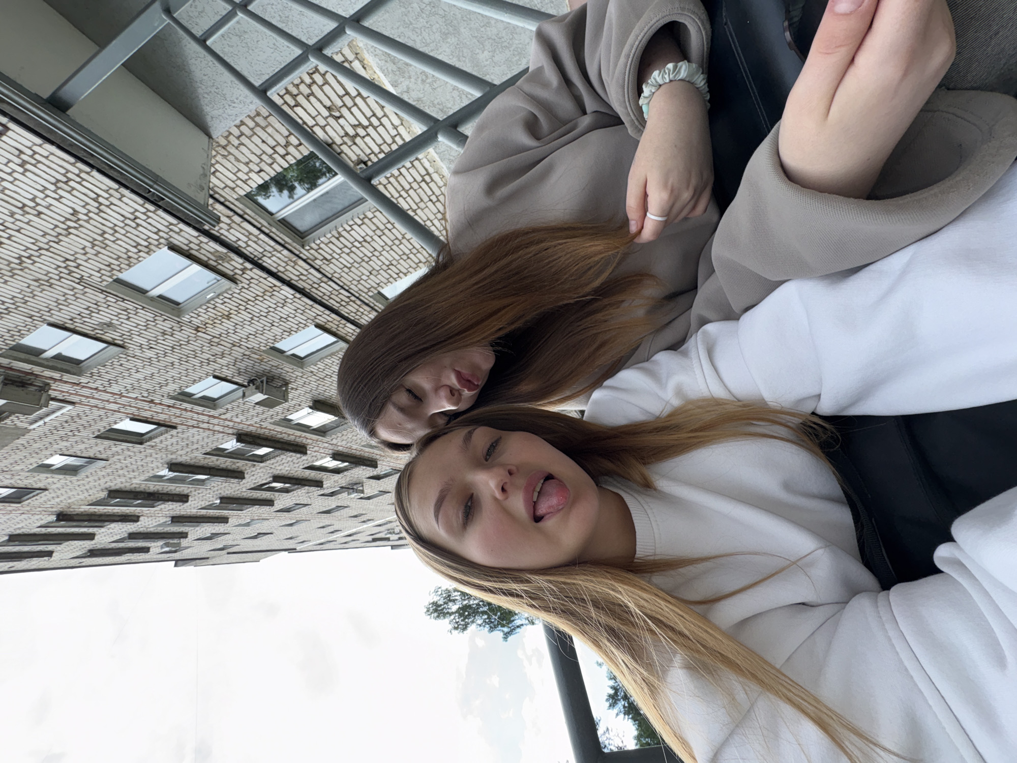Two young females are posing for a selfie outdoors, in front of a brick apartment building. The building appears to be a multi-story structure with numerous windows. A metal railing is visible to the right of the individuals. The sky is overcast. The female in the foreground, on the left, is leaning her head slightly to the side and sticking her tongue out. She has long blonde hair and is wearing a white sweatshirt. The female behind her, on the right, has her eyes closed and her lips pursed, as if she is blowing a kiss or making a duck face. She has long brown hair and is wearing a beige sweatshirt. The specific location is identified as being in the city of Chaikovsky, Russia.