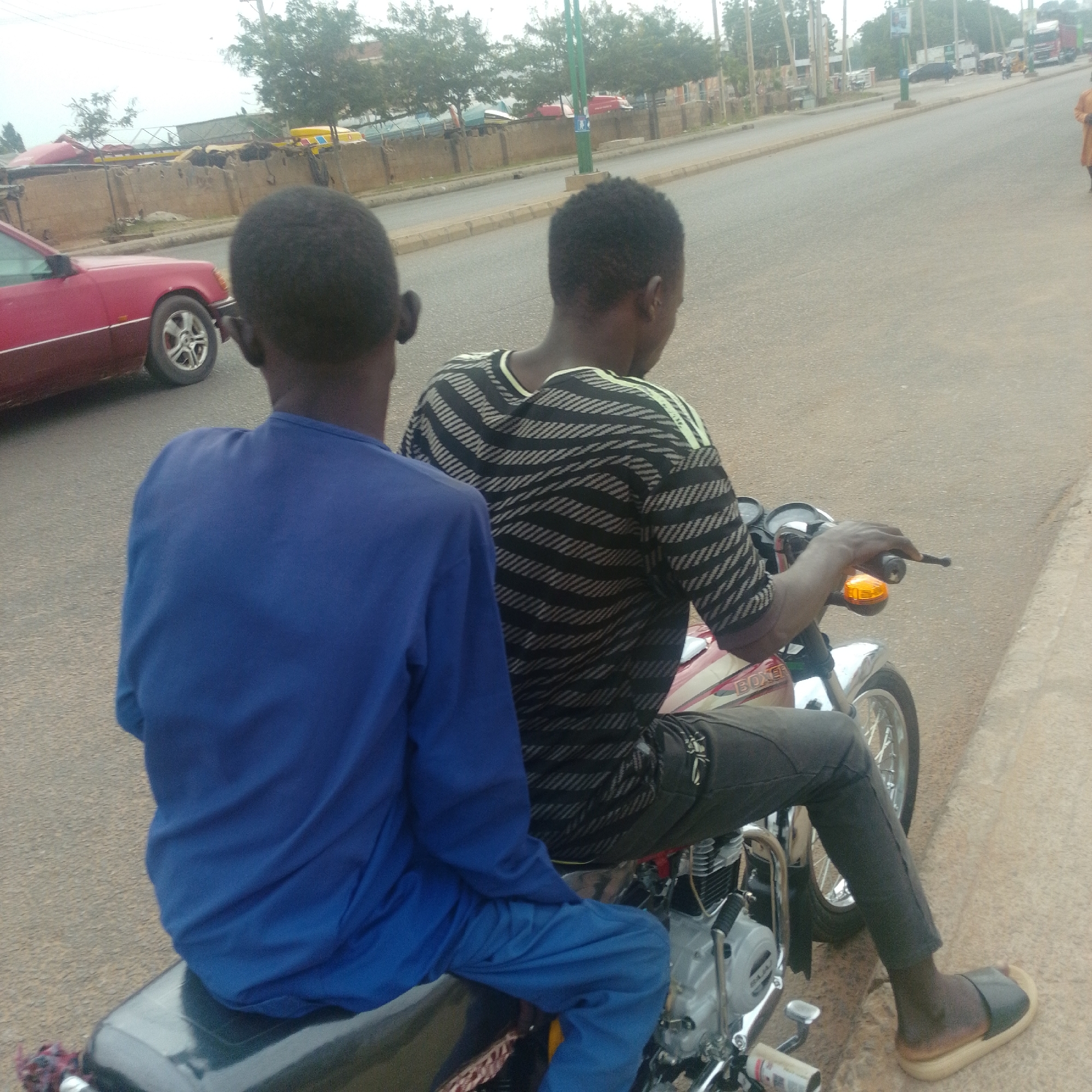 Two young males are on a motorcycle on a paved road in Gombe, Nigeria. The male in the foreground is wearing a blue long-sleeved shirt and blue pants. He is facing away from the camera, seated on the back of the motorcycle. The male in the background is wearing a black and white striped shirt and grey pants. He is seated in front of the other male, driving the motorcycle. The motorcycle is red and white with "BOXER" written on its side. A red car is parked on the left side of the road. The environment is urban, with trees and buildings visible in the background. The sky is overcast. The road appears to be a main thoroughfare, with other vehicles and pedestrians potentially present out of frame.