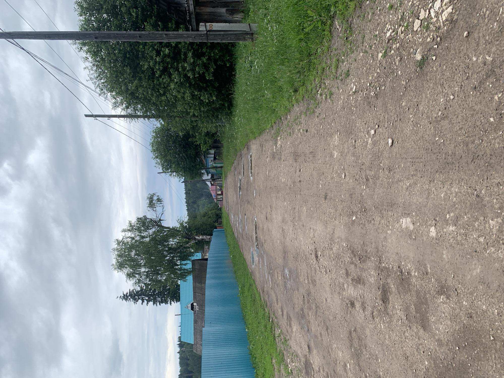 The image depicts a dirt road in a rural area, likely a village, with houses and trees lining its sides. The road surface is uneven and shows signs of recent rain, with several puddles visible. A bright turquoise fence runs along the left side of the road, bordering a property with a house featuring a blue roof. On the right side of the road, there is a wooden utility pole with electrical wires running overhead, and an old wooden structure. The sky is overcast, suggesting a cloudy day. The background shows a dense forest and rolling hills. The scene appears to be in a tranquil, undeveloped part of Barsai, Russia.